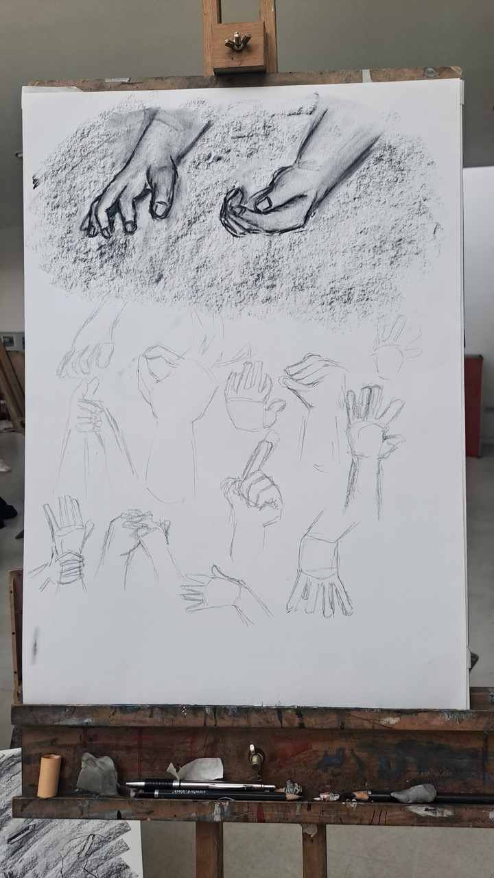 A sketch on a white canvas shows a pair of hands drawn in black pencil at the top, with lighter hand studies below on an easel in a studio.