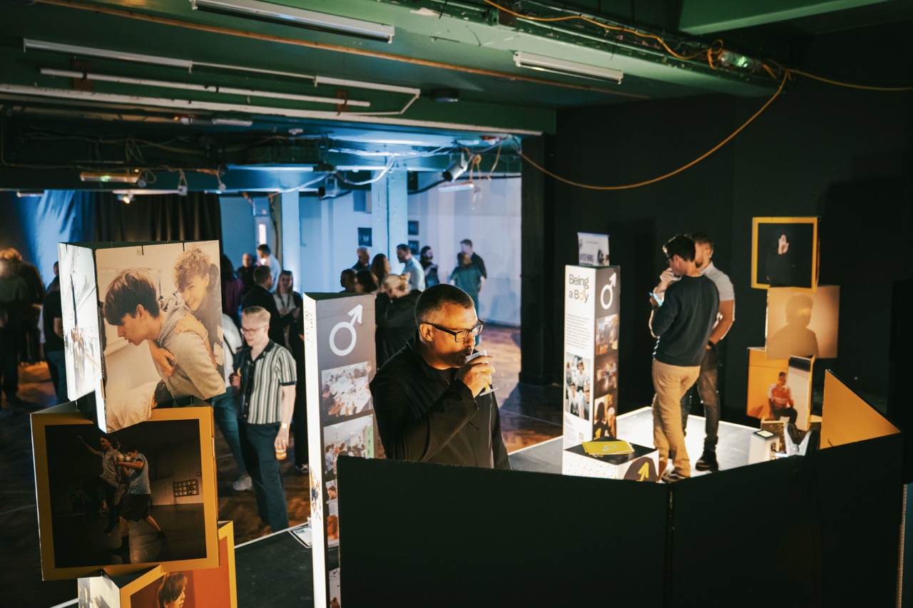 People viewing photographic displays in a dimly lit exhibition space, with framed images and exhibition panels arranged between black partitions.