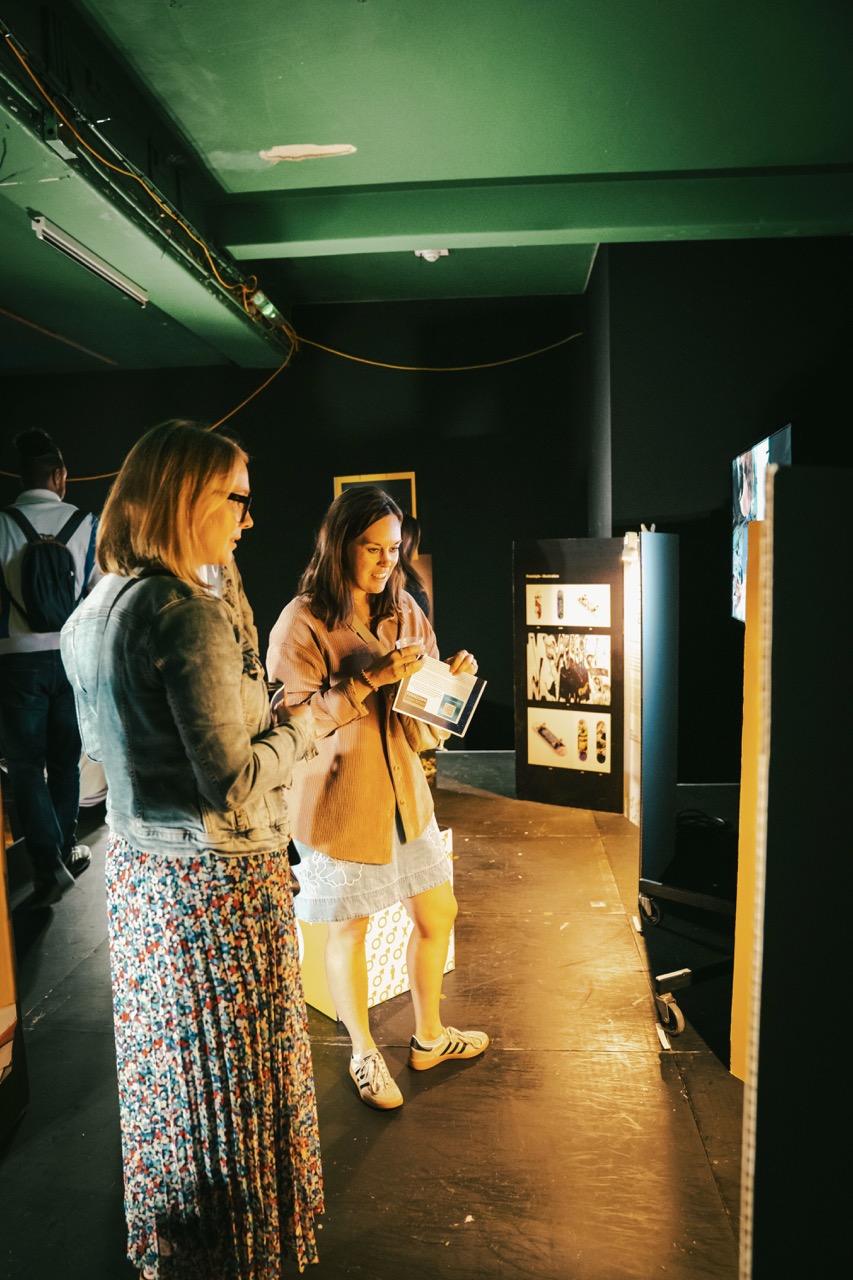 Two women stand in a dimly lit exhibition space, looking at a display board and holding a booklet.