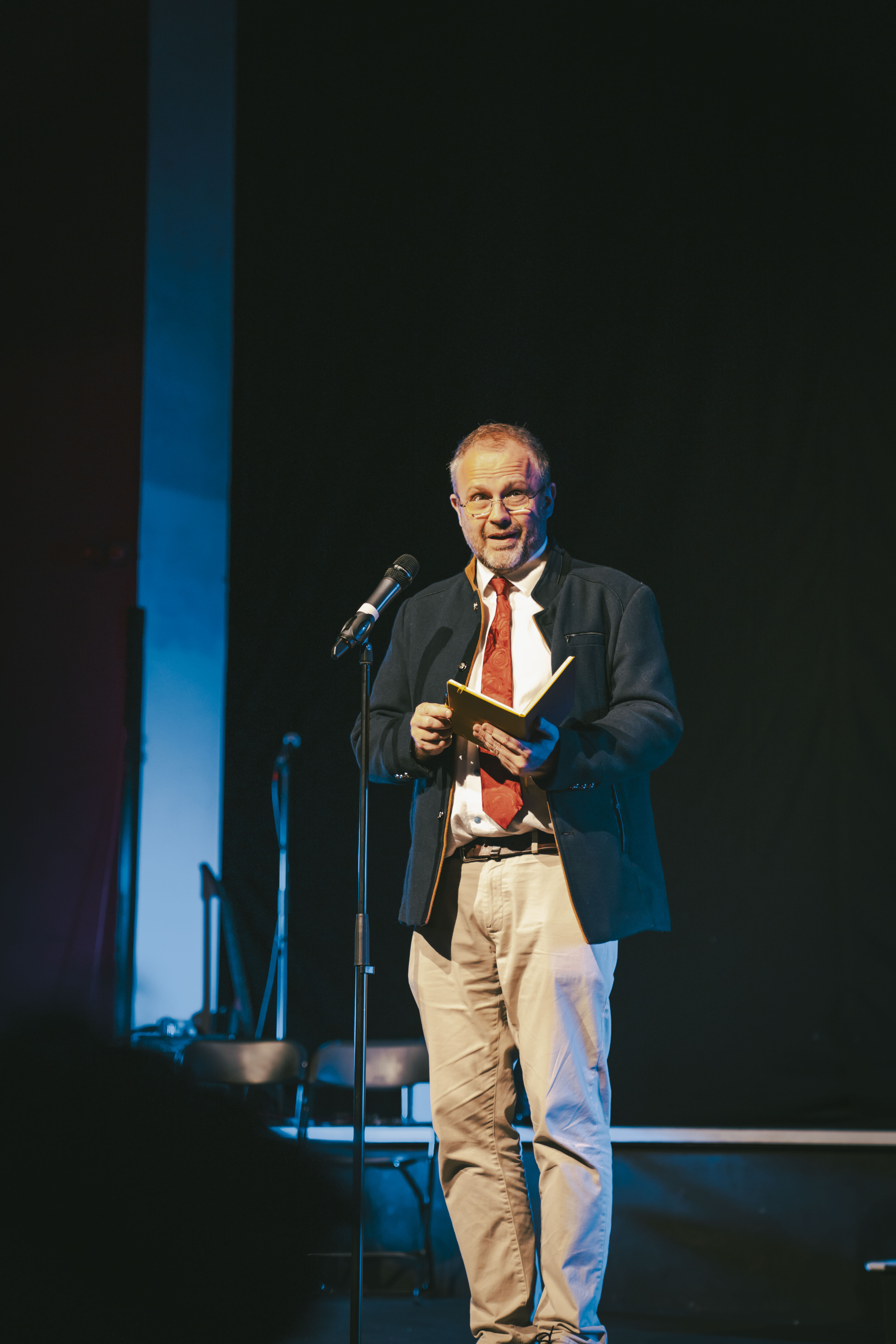An older man in a blazer and red tie stands on a stage holding a book beside a microphone stand, with dark lighting and chairs in the background.