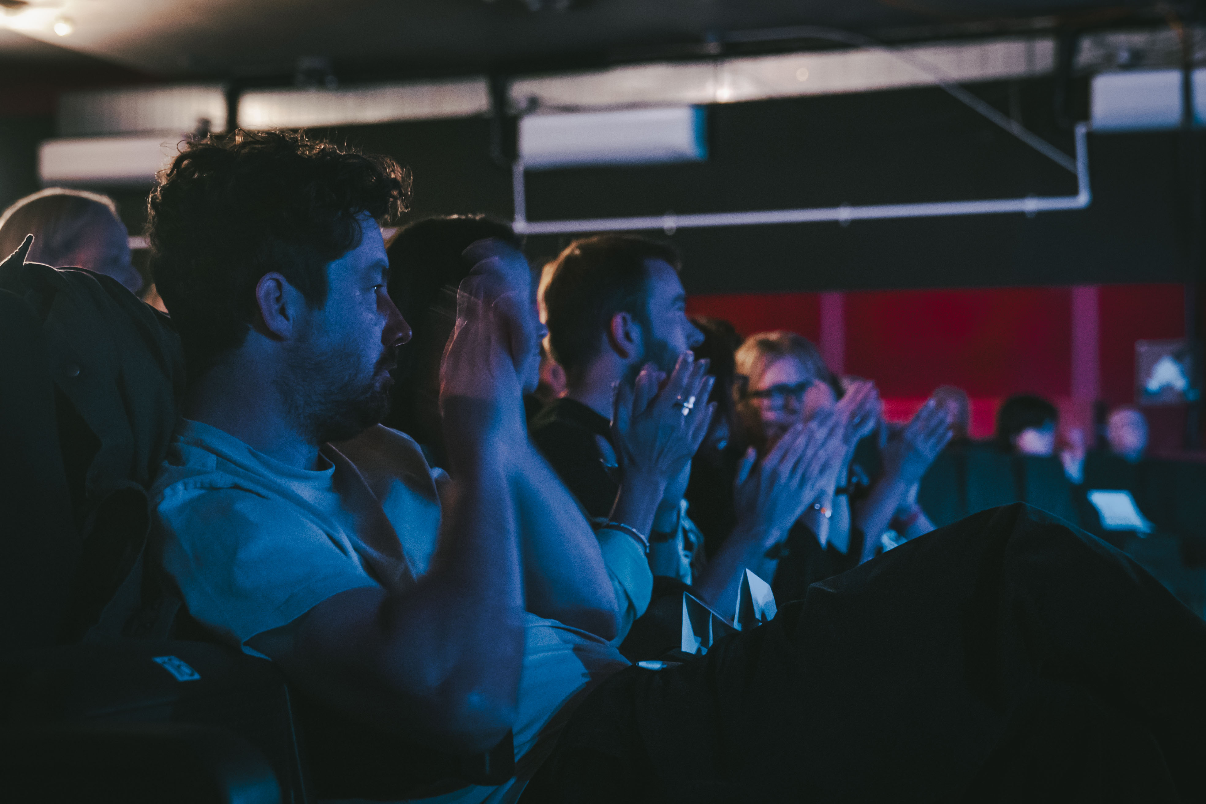 People seated in an audience, applauding under blue and red stage lighting in a dark venue.