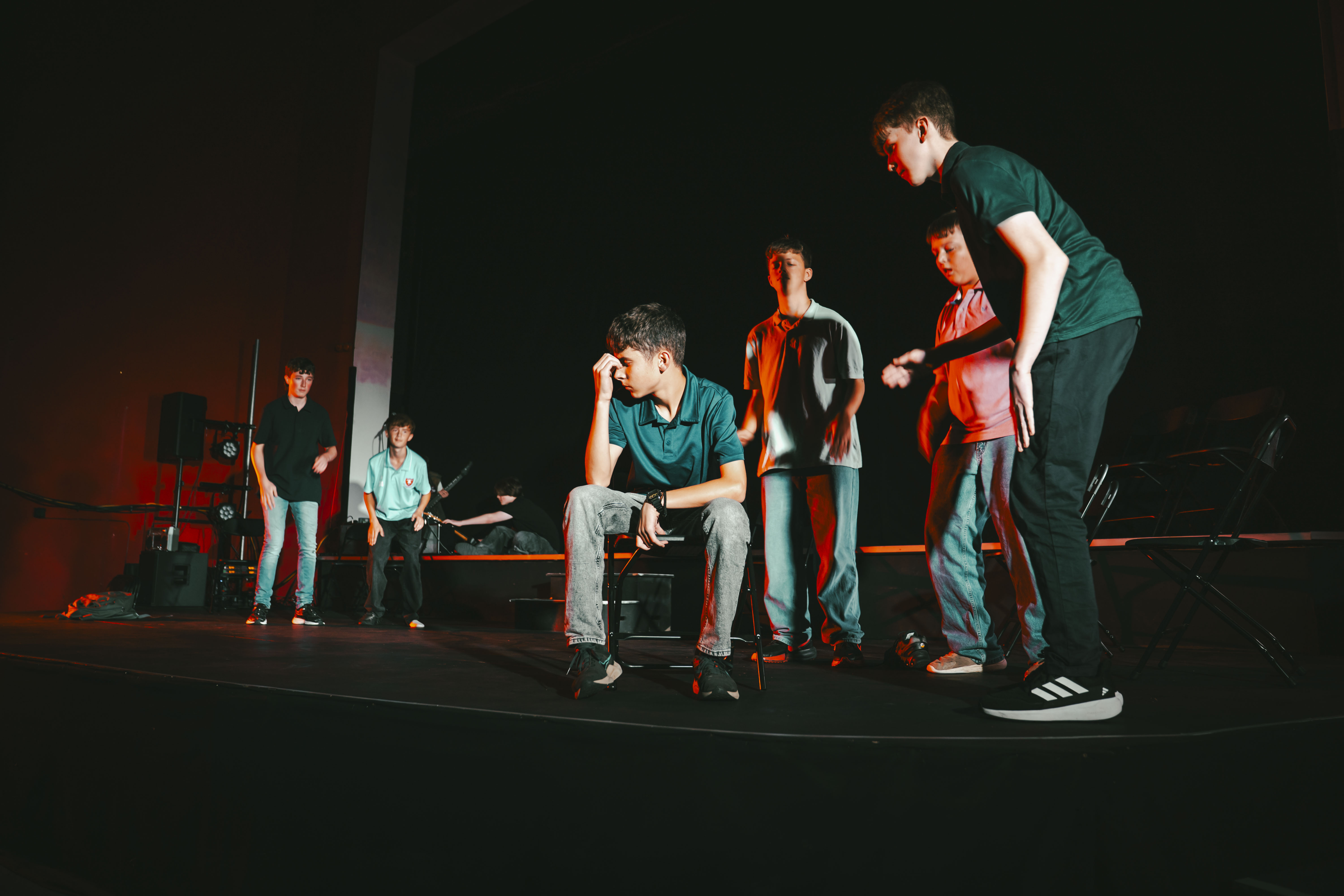 Five young men stand and sit on a dark stage with red lighting, with a drum kit and speakers in the background.