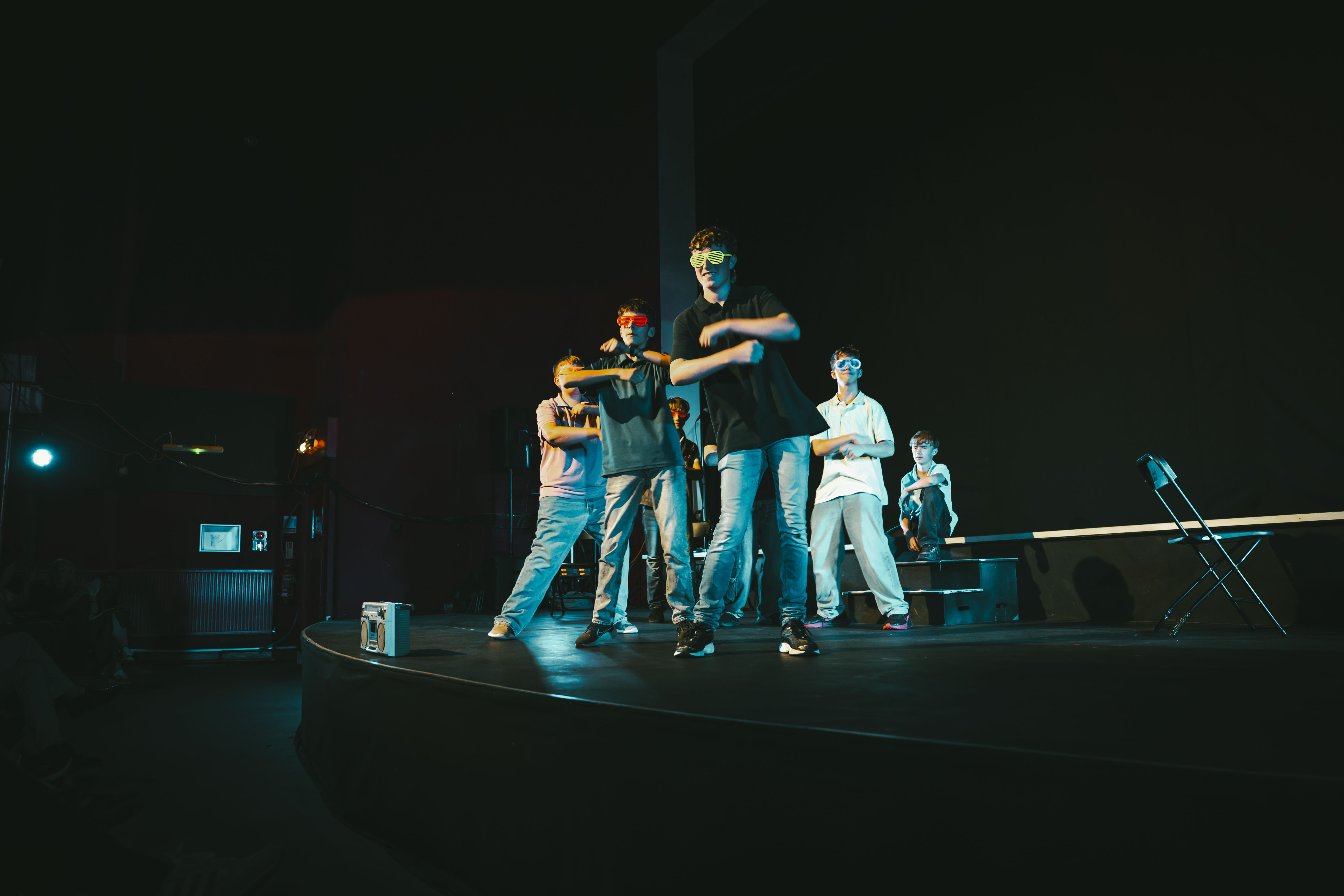 Five men in casual clothes perform a choreographed dance on a dark stage with blue lighting, while a stool and chair sit at the side.