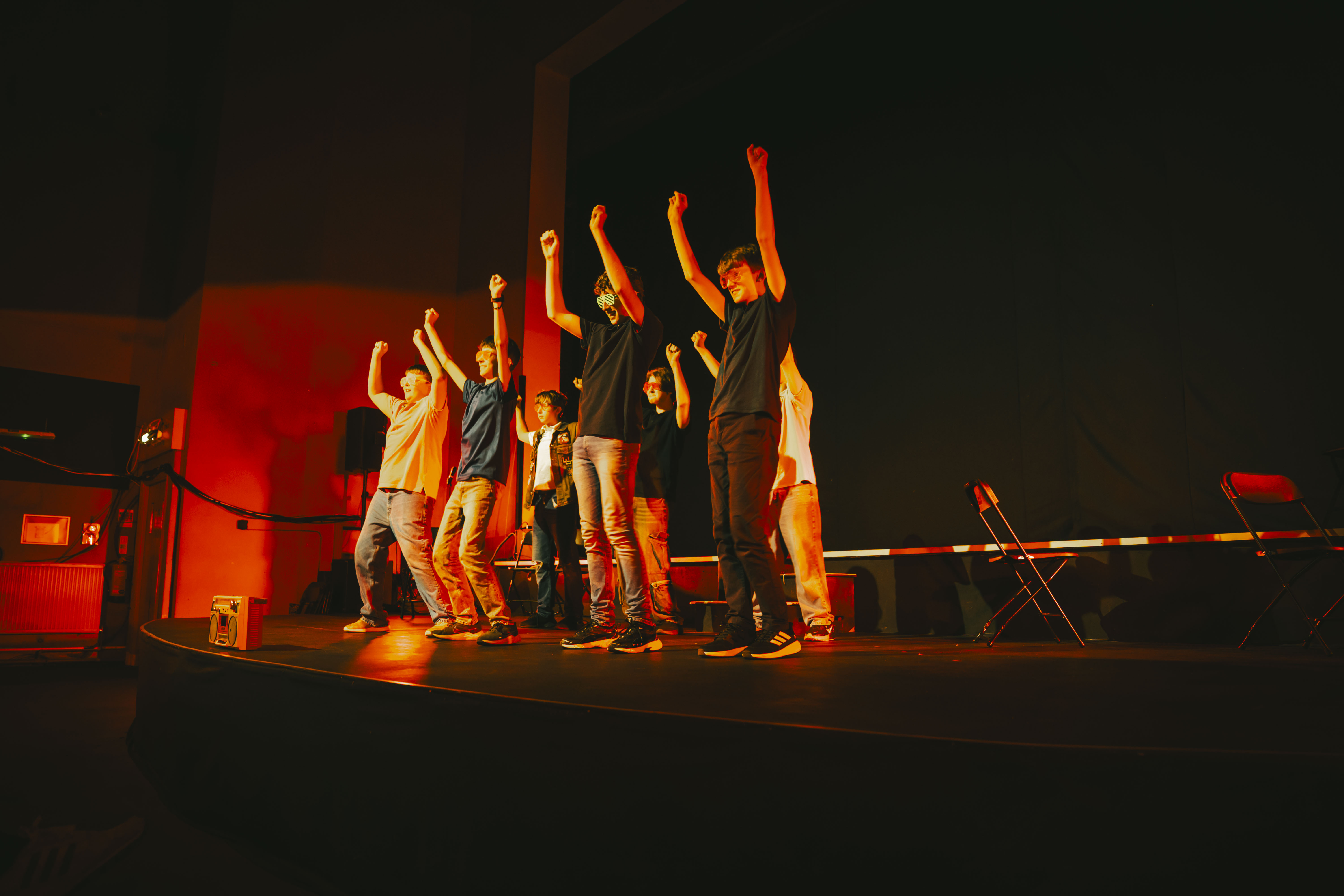 A group of people on a dark stage with both arms raised, lit by red and orange lighting, with a keyboard stand in the background.