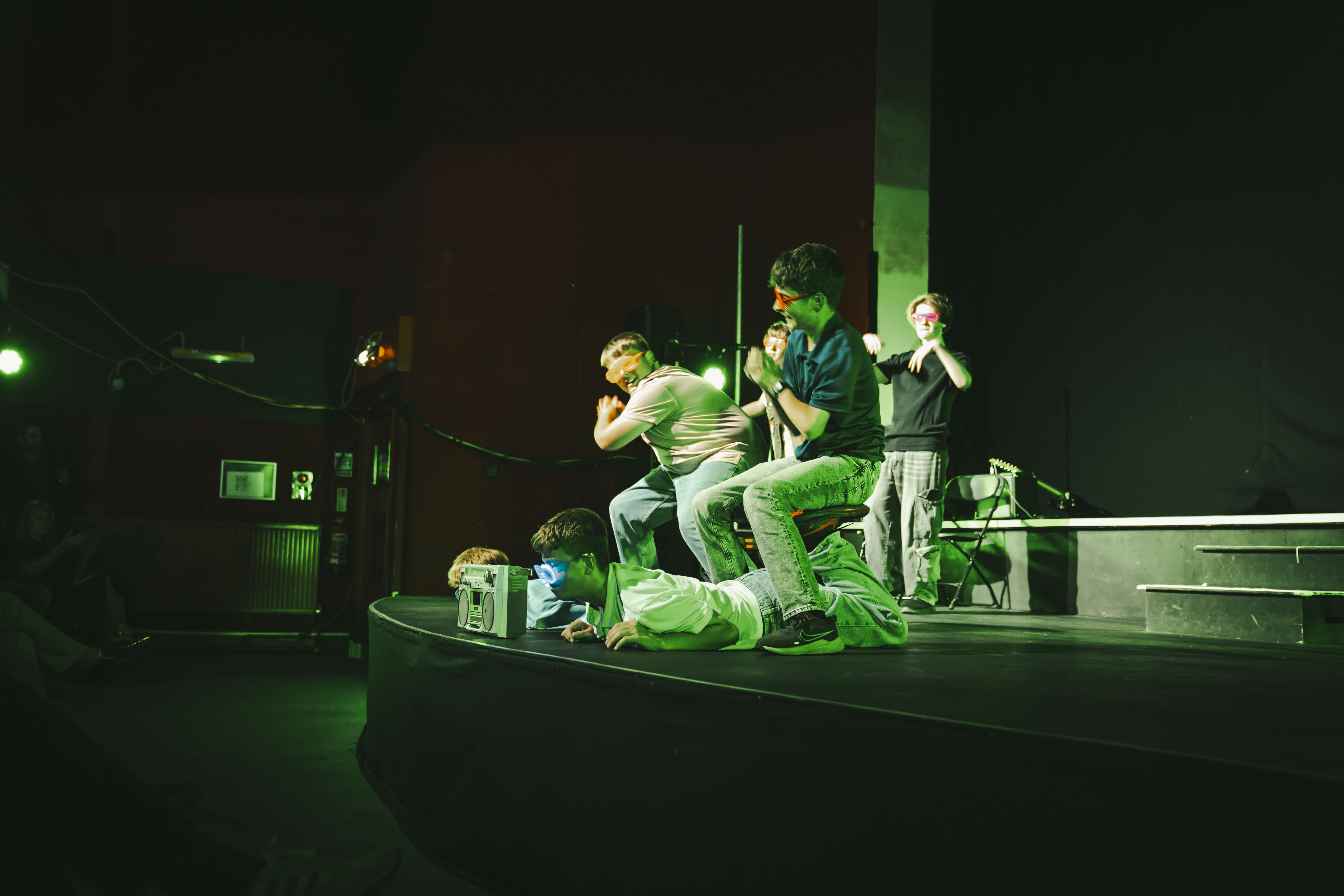 Three people performing on a dark stage under green lighting, with one person kneeling in the foreground and two others standing behind near a raised platform.