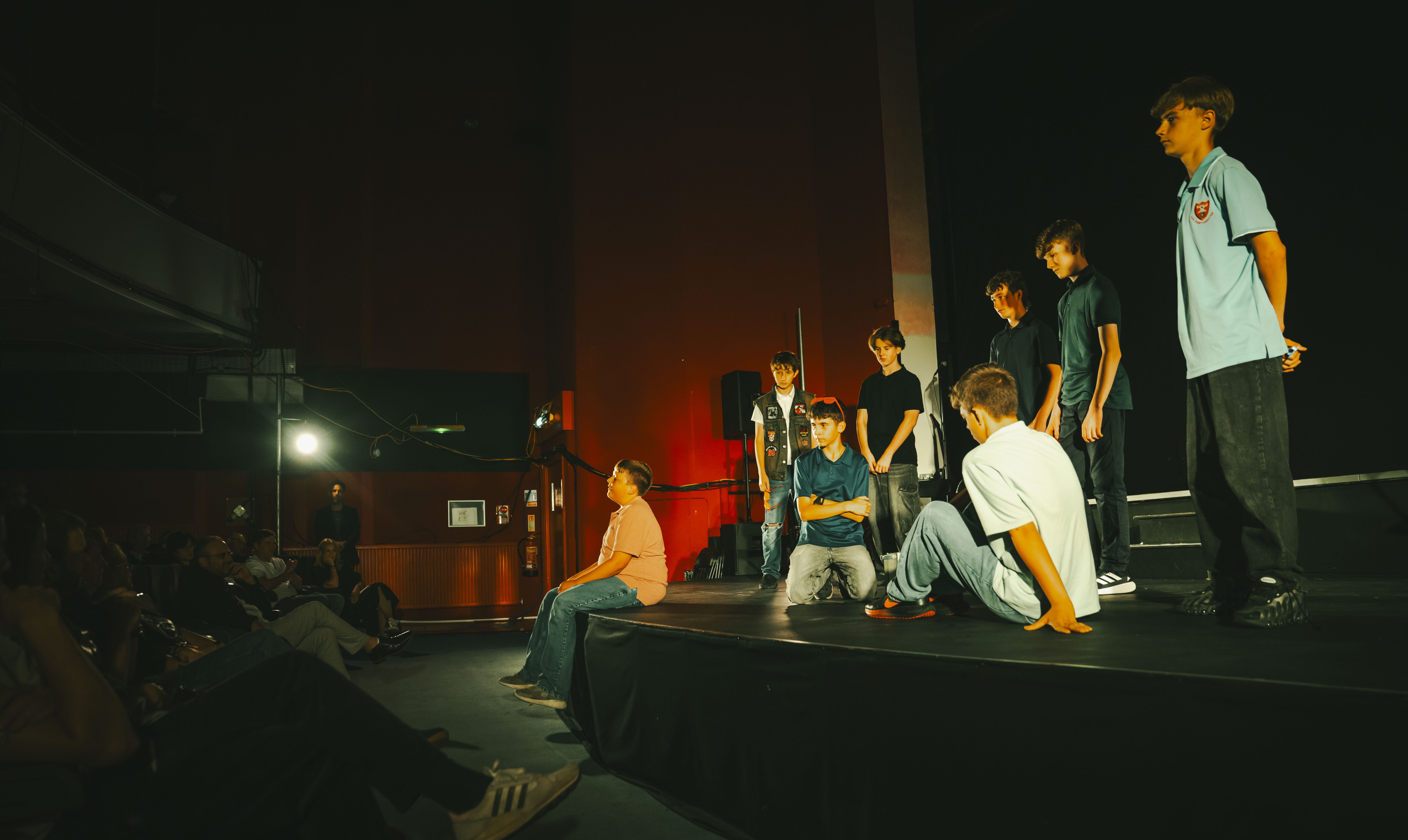 A group of children and teenagers on a low stage in a dark indoor venue, with seated audience members in the foreground and red lighting on the back wall.