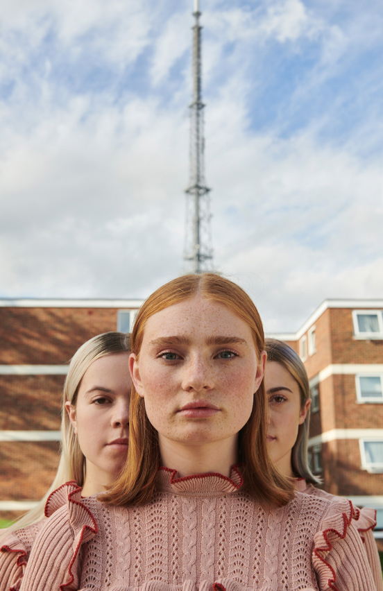 Photo of three people facing forward, standing in front of a block of flats with a metal tower in the background.