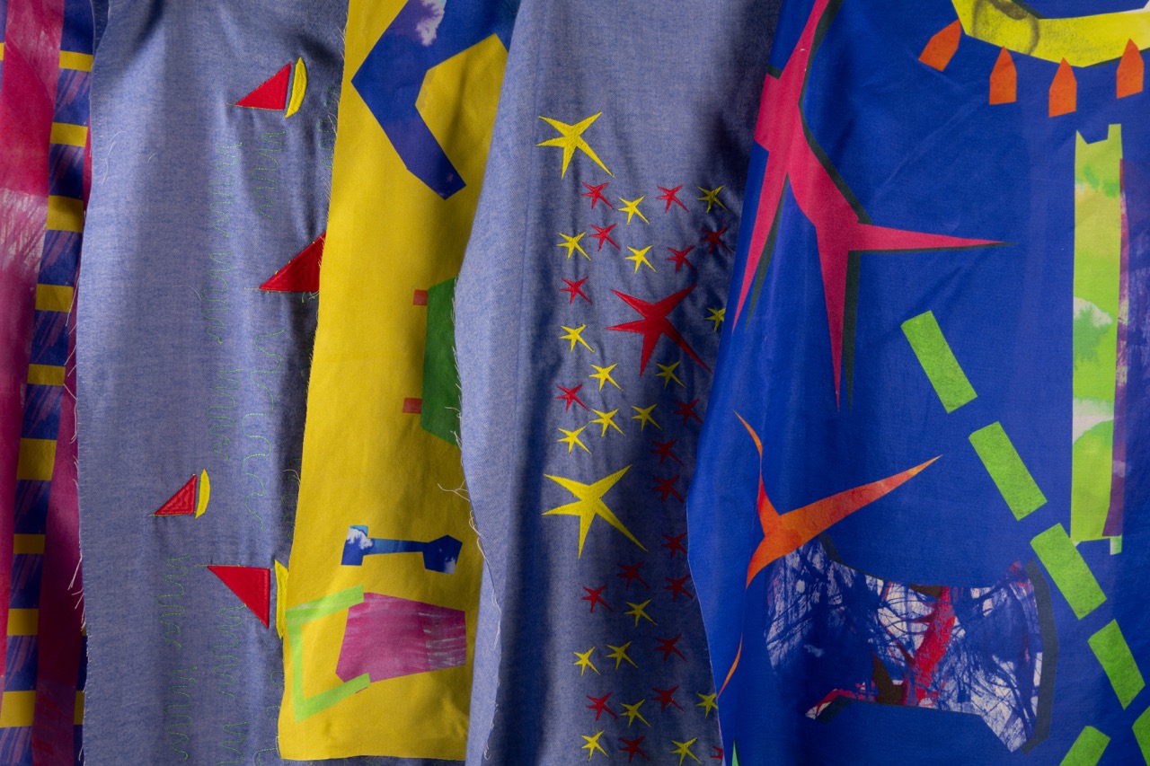 Five pieces of fabric hanging side by side, featuring a range of vibrant colours and geometric patterns. The fabrics include yellow, blue, and red segments with abstract shapes, stars, and various cutouts, creating a visually dynamic display.