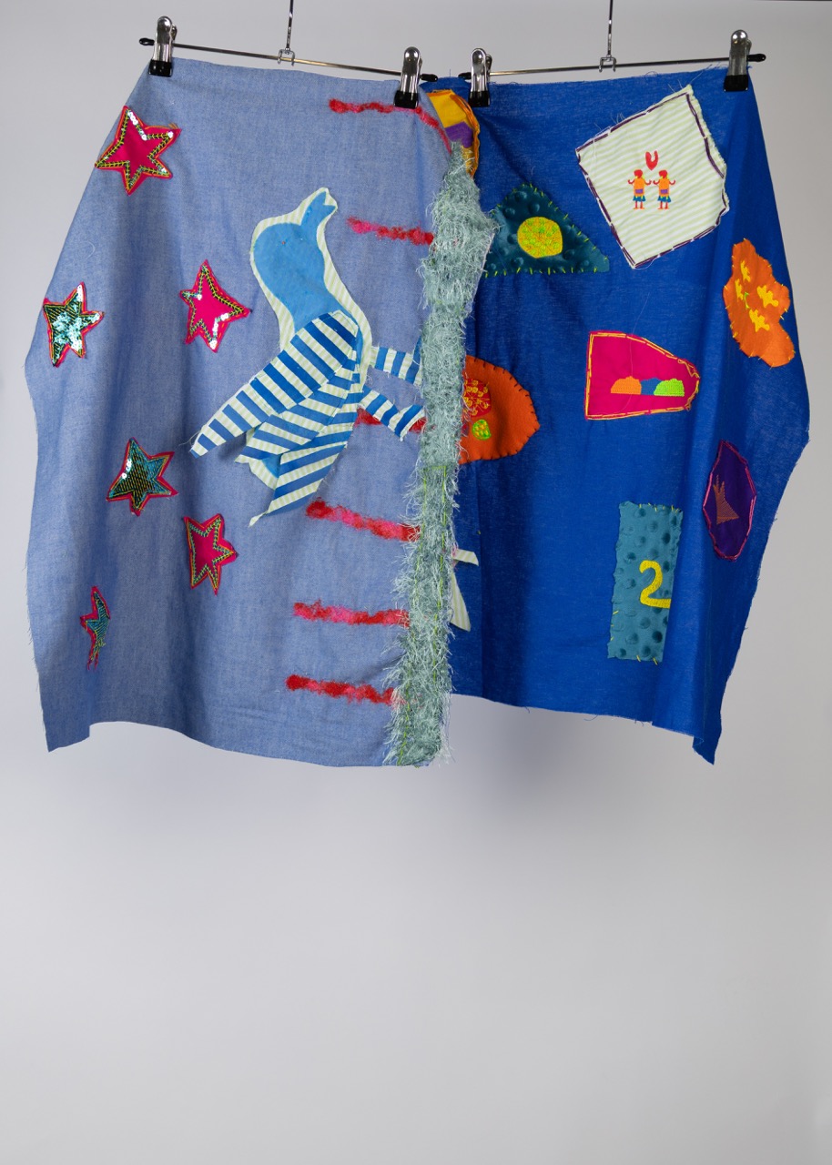 A blue fabric skirt displayed on a hanger, featuring various decorations including colorful stars, a striped figure, and abstract shapes in orange, green, and yellow. The skirt includes attached textures and patterns, giving it a vibrant, artistic appearance.