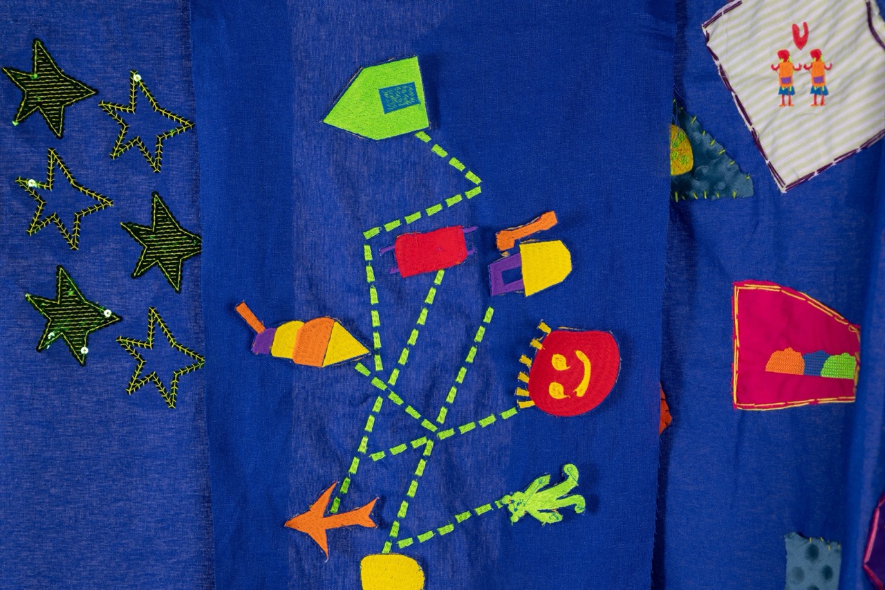 A blue fabric showing colourful appliqués including stars, geometric shapes, and a playful character with a smiling face. The designs are connected by dashed lines, creating a whimsical scene across the textile.