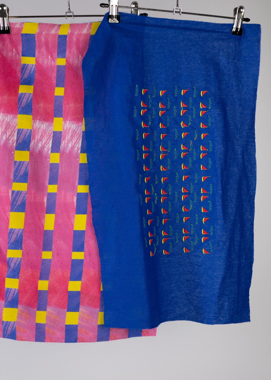 Two pieces of fabric displayed on clips against a white background. The left fabric features vibrant pink and yellow patterns in a grid layout, while the right fabric is a solid blue with a smaller, repeated geometric design in various colours.