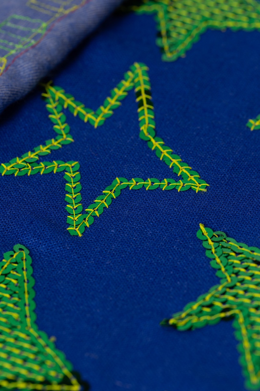 A close-up of blue fabric featuring yellow and green star-shaped embroidery. The stitching is visible, with a detailed pattern creating contrasting star designs on the textured surface.