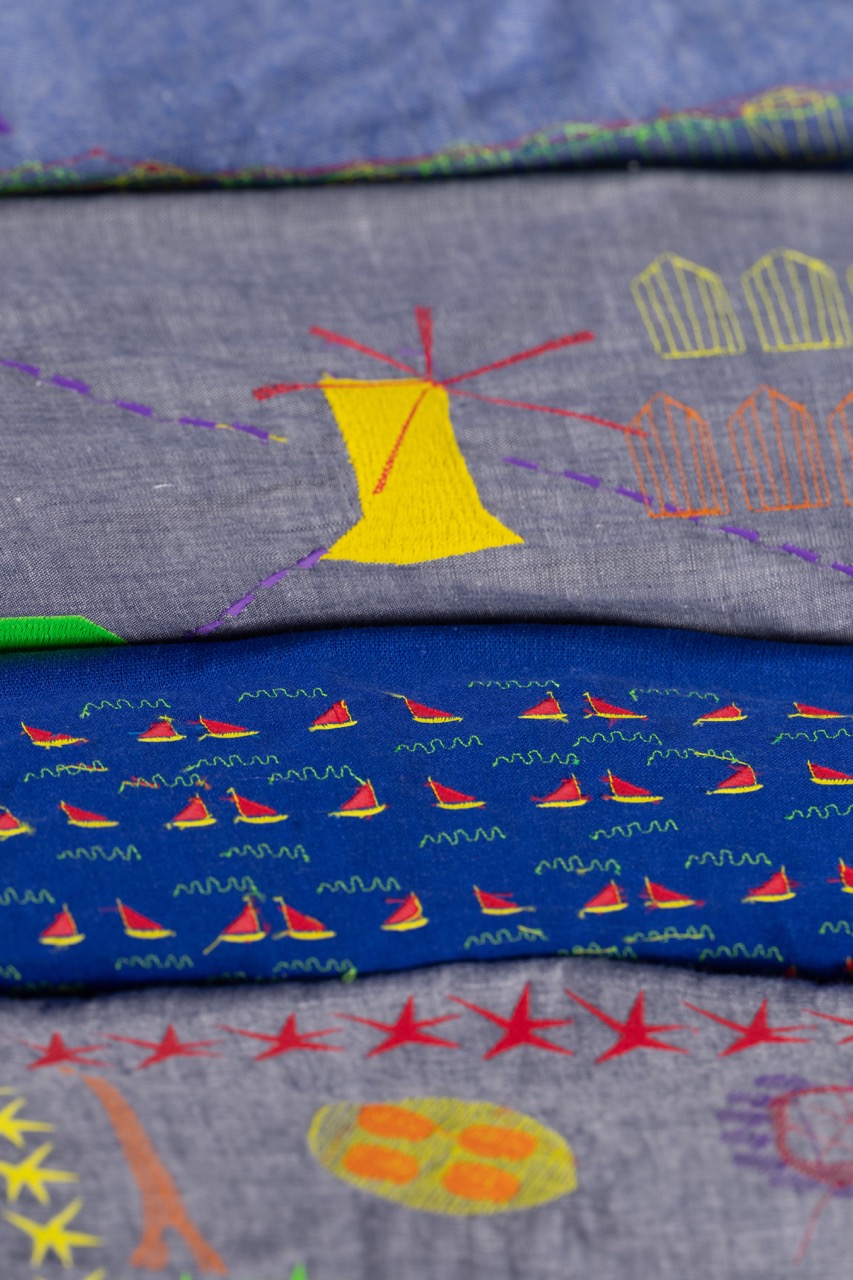 A stack of folded fabrics featuring colourful embroidery. The top layer has a grey background with various green and yellow designs, the second shows a yellow windmill, and the bottom layer depicts blue water with red boats and green accents.