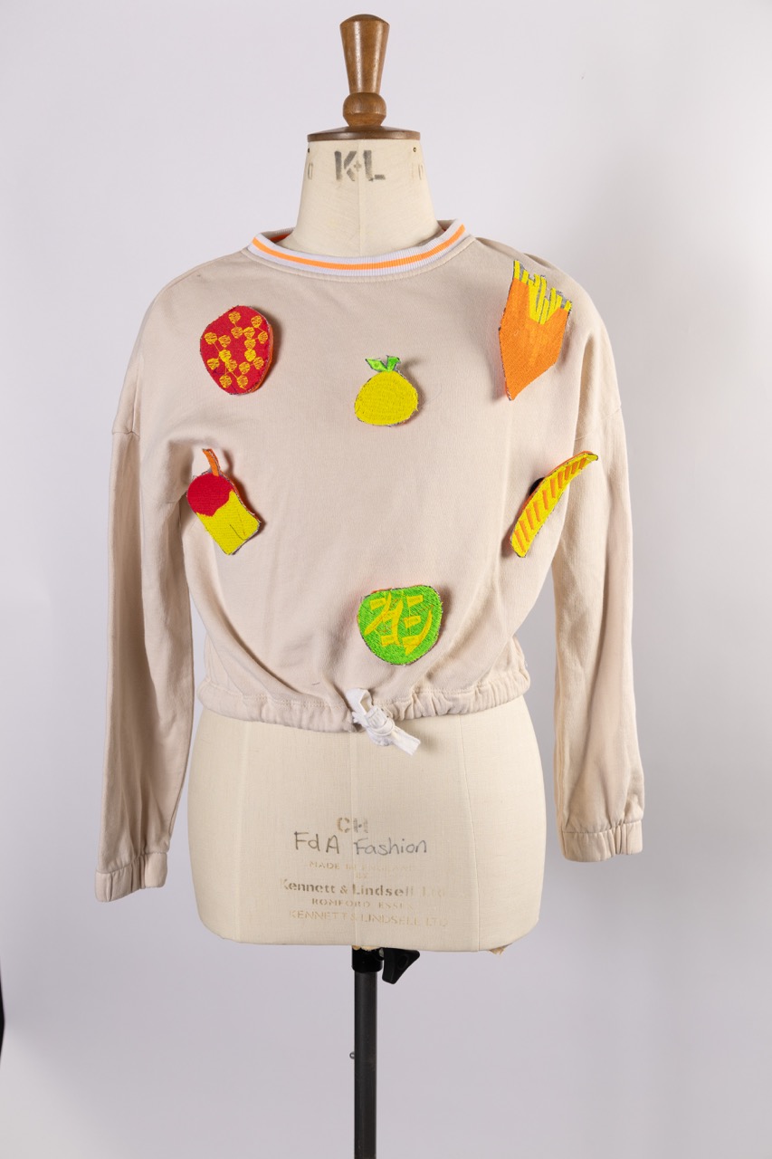 A beige sweatshirt displayed on a dress form features colourful appliqué designs, including a pineapple, a carrot, a red cup, and other fruit and vegetable shapes, with a gathered hemline and striped collar.
