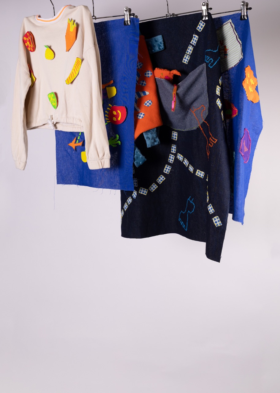 A display of various fabric pieces and a sweater hanging on clips. The sweater features colourful fruit and vegetable appliqués. The fabrics include a blue piece with patterned designs and a dark fabric with embroidered details.