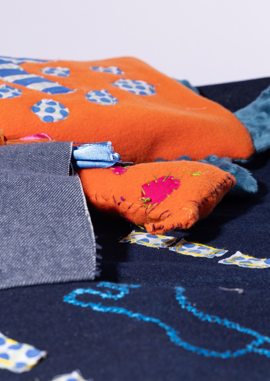 An orange and navy textile piece with vibrant decorations, including geometric patterns and floral embroidery. The piece is layered with a denim-like fabric, showcasing an artistic design and textural contrast.