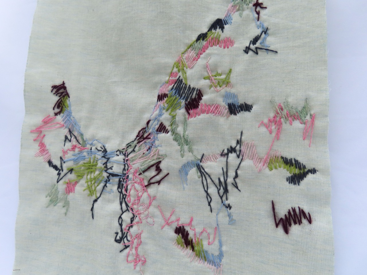A piece of cream fabric featuring various abstract embroidery patterns in multiple colours, including pink, green, blue, and black threads. The stitching forms loose, intertwining shapes and lines, creating an artistic design.