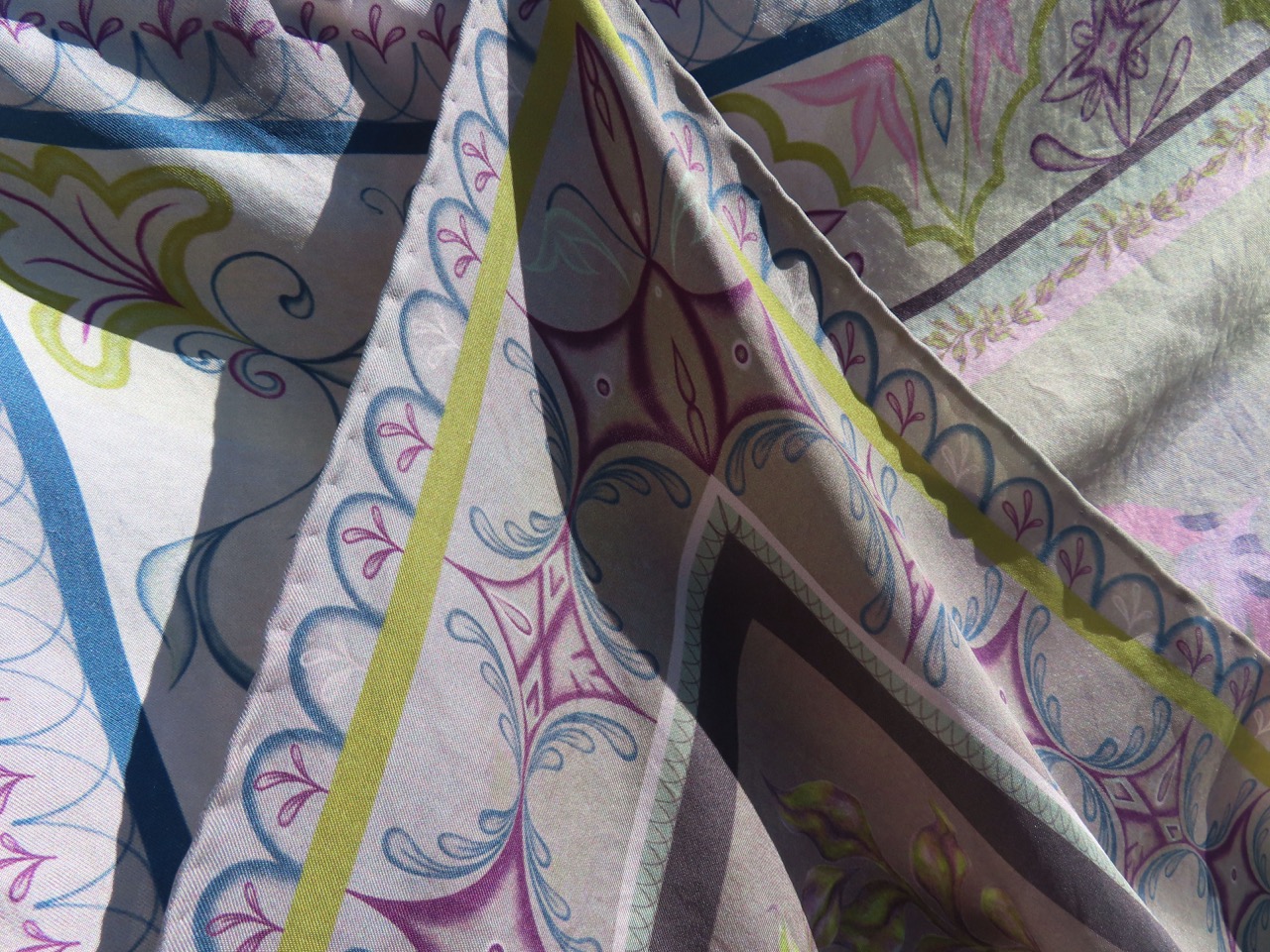 A close-up of a patterned fabric displaying intricate designs in pastel colours. The fabric features floral motifs in pink, blue, and green, with swirls and geometric lines, creating a visually appealing texture.