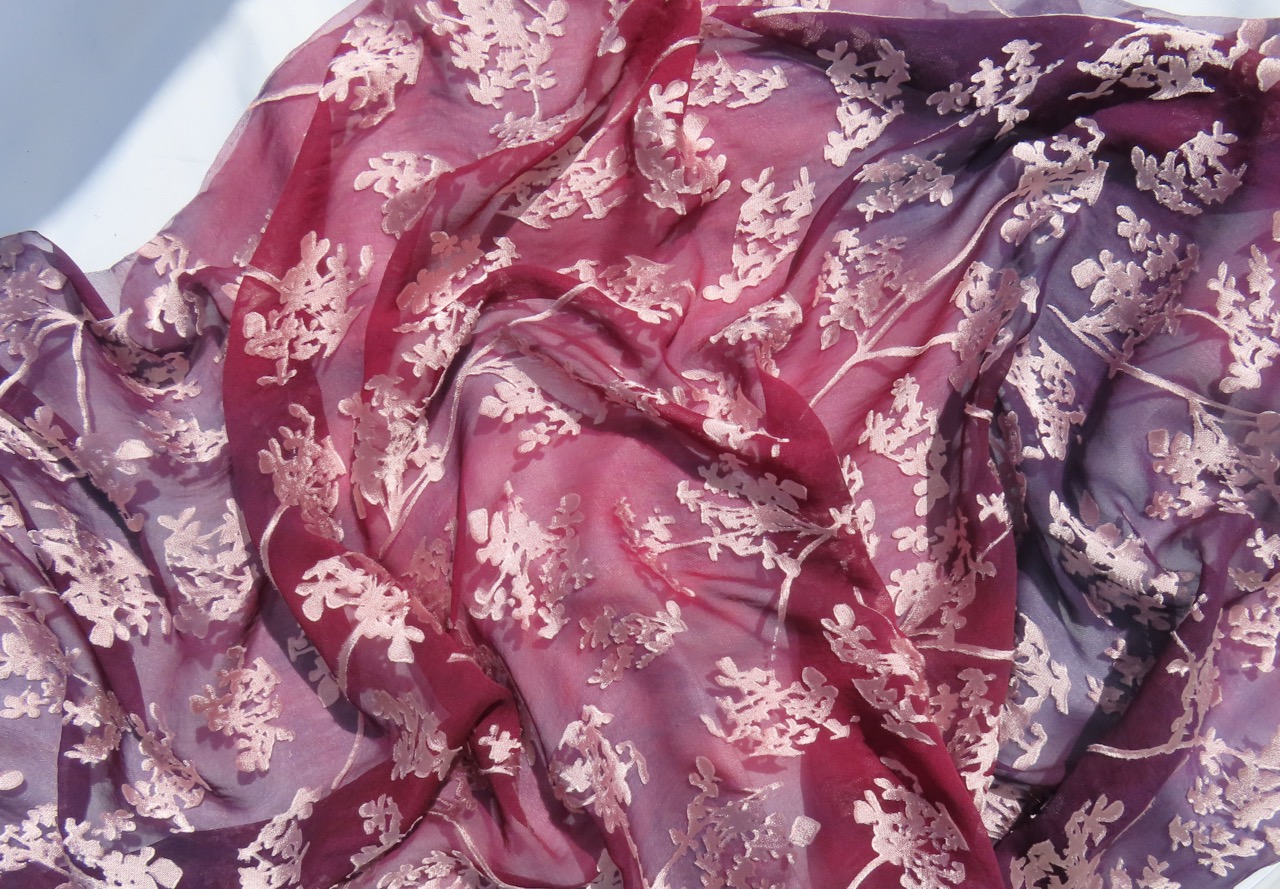 A flowing piece of fabric in shades of deep red and purple, adorned with delicate floral patterns in pale pink. The silk appears lightweight and translucent, with softly draped folds creating a textured surface.