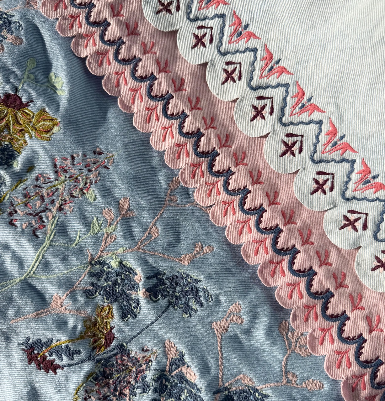 A close-up of fabric featuring intricate embroidery. The left side shows blue fabric with floral patterns in shades of purple, pink, and yellow, while the right side displays a light fabric with scalloped edges and pink and red embroidery patterns.
