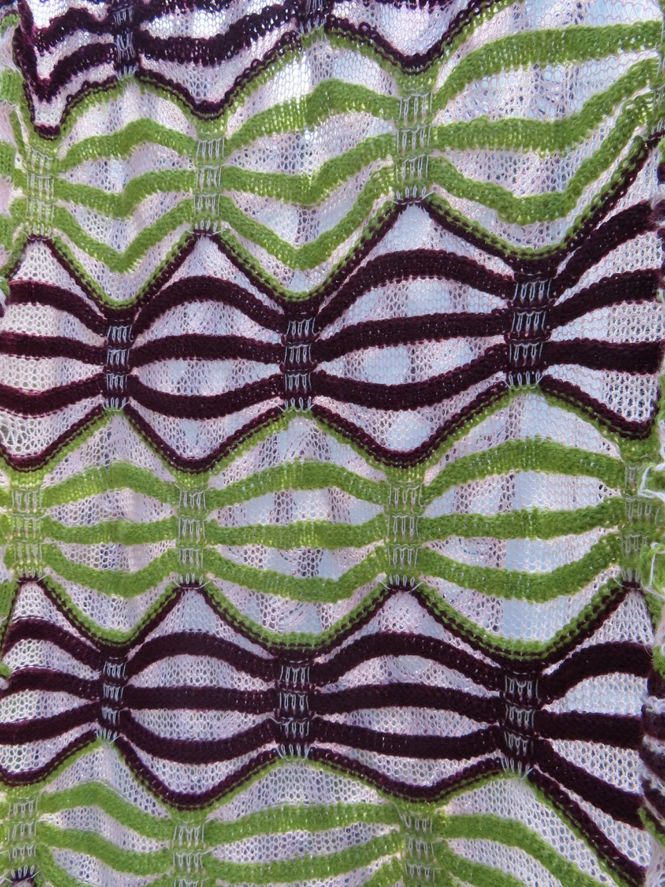 A textured fabric featuring a repeating zigzag pattern with alternating sections of dark brown, green, and white. The design creates a visual contrast with waves formed by the stripes.
