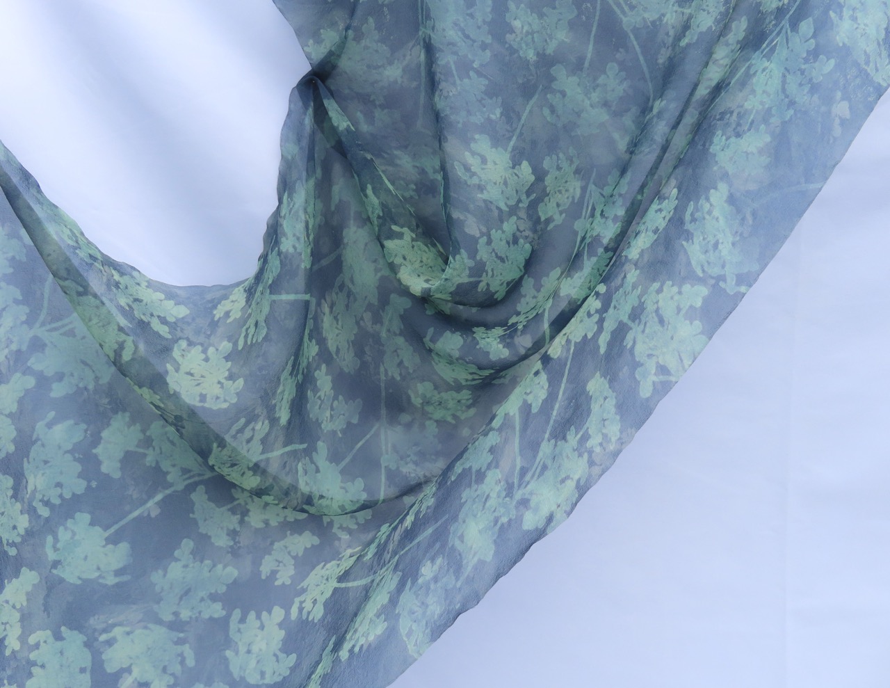 A sheer scarf featuring a leafy design in green tones against a translucent background. The fabric drapes softly, creating gentle folds and revealing the pattern clearly.