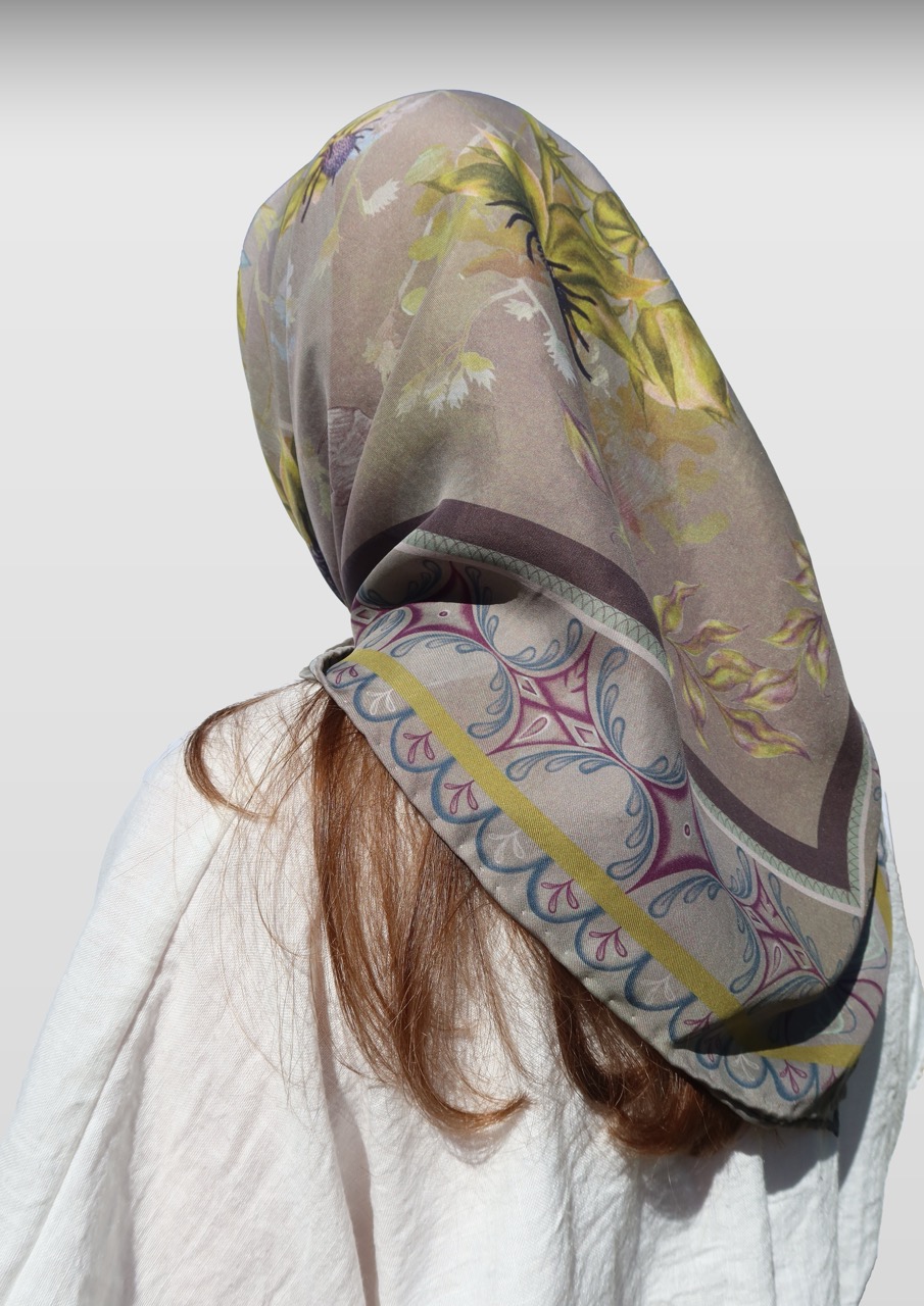 A person with long brown hair, partially visible from the back, wears a large patterned scarf draped over their head and shoulders. The scarf features floral designs and decorative borders on a muted background.