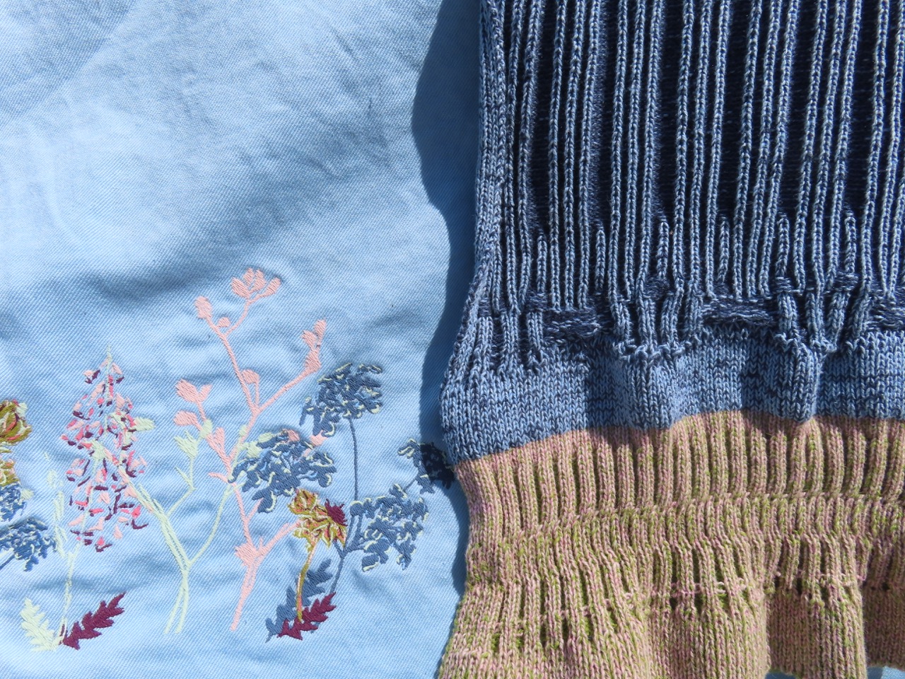 A close-up of two fabrics: on the left, a light blue textile with colourful floral embroidery featuring pink, yellow, and blue designs; on the right, a knitted fabric in blue and tan with ribbed and patterned sections.