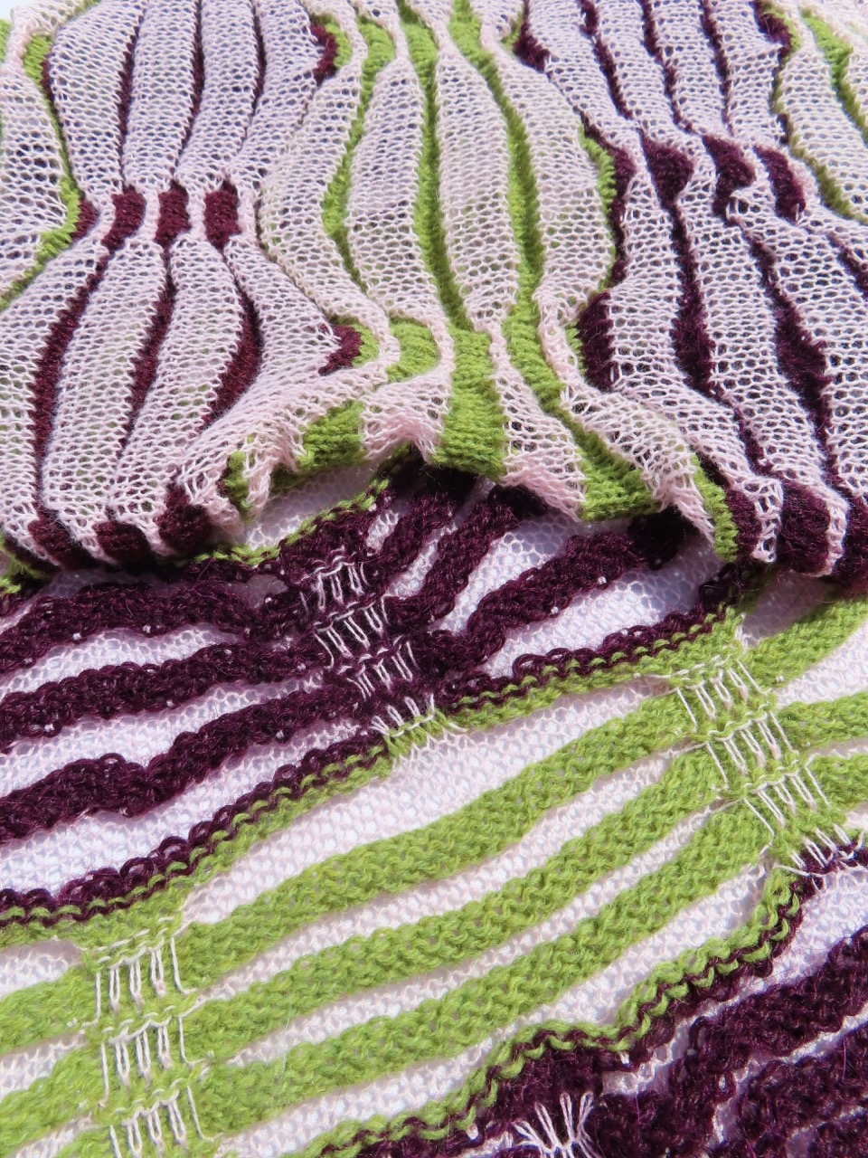 A close-up of knitted textile featuring an intricate pattern in shades of green, cream, and deep purple. The fabric showcases wavy lines and varying textures, highlighting the craftsmanship in the design.