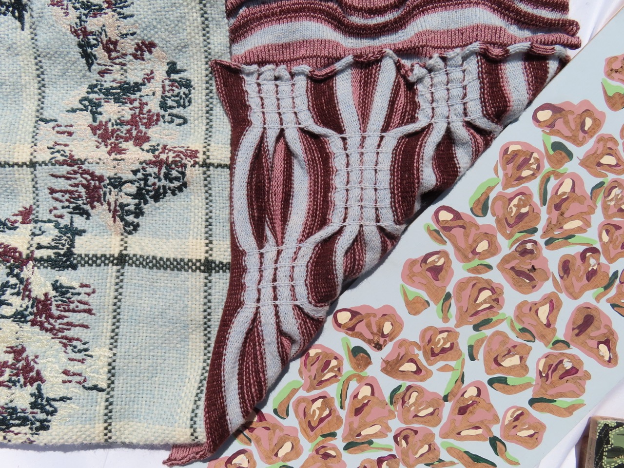 A close-up of assorted textiles, including a patterned fabric featuring floral designs, a striped knit fabric in maroon and grey, and a textured fabric with a checkered pattern in muted colours. The arrangement showcases diverse textures and patterns.
