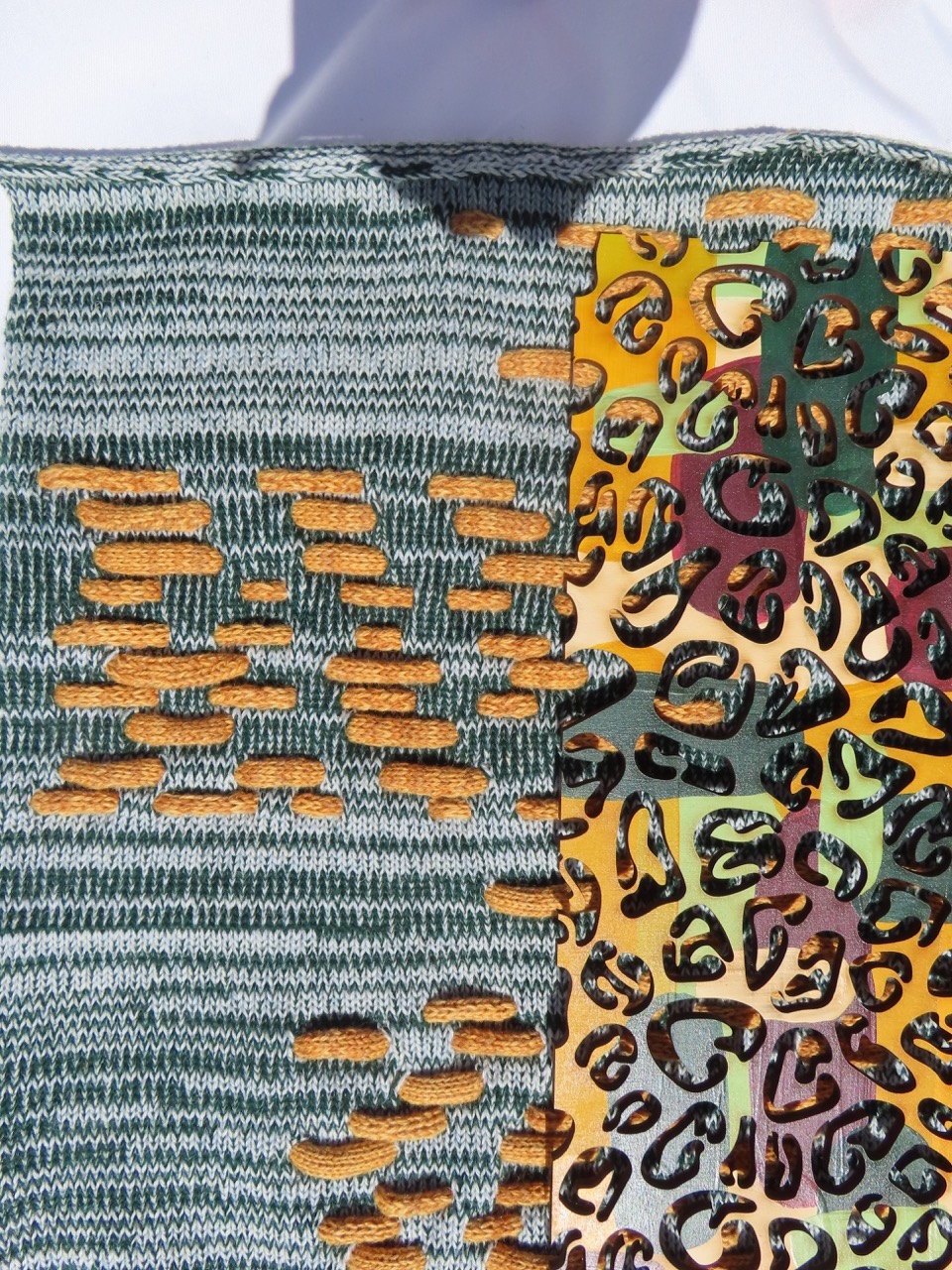 Fabric with a textured green knitted section featuring gold and tan patterns interspersed with patches of various colours. A segment on the right displays a vibrant, animal-print design in shades of yellow, green, and brown, adding contrast to the woven texture.