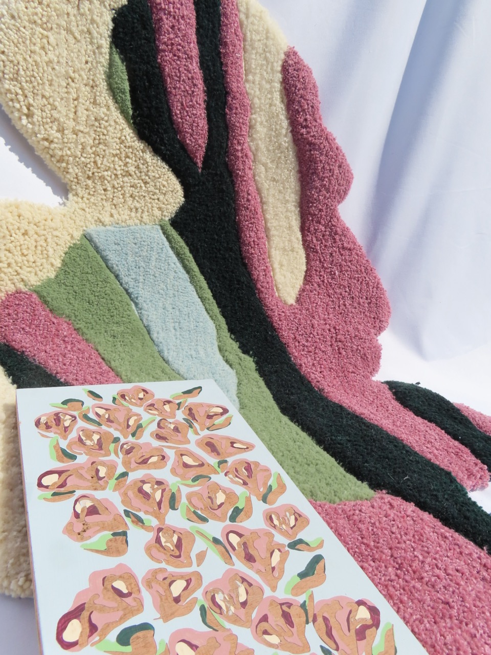 A textured rug with abstract shapes in shades of pink, green, black, and cream, positioned beside a flat surface displaying a sheet of patterned paper featuring several pink flowers on a pale blue background.