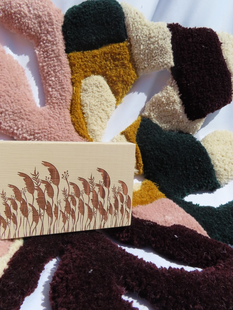 A rectangular box with a simple design featuring brown foliage illustrations is positioned in front of an abstract, colourful textile background made of shaggy materials in shades of pink, green, yellow, and burgundy.