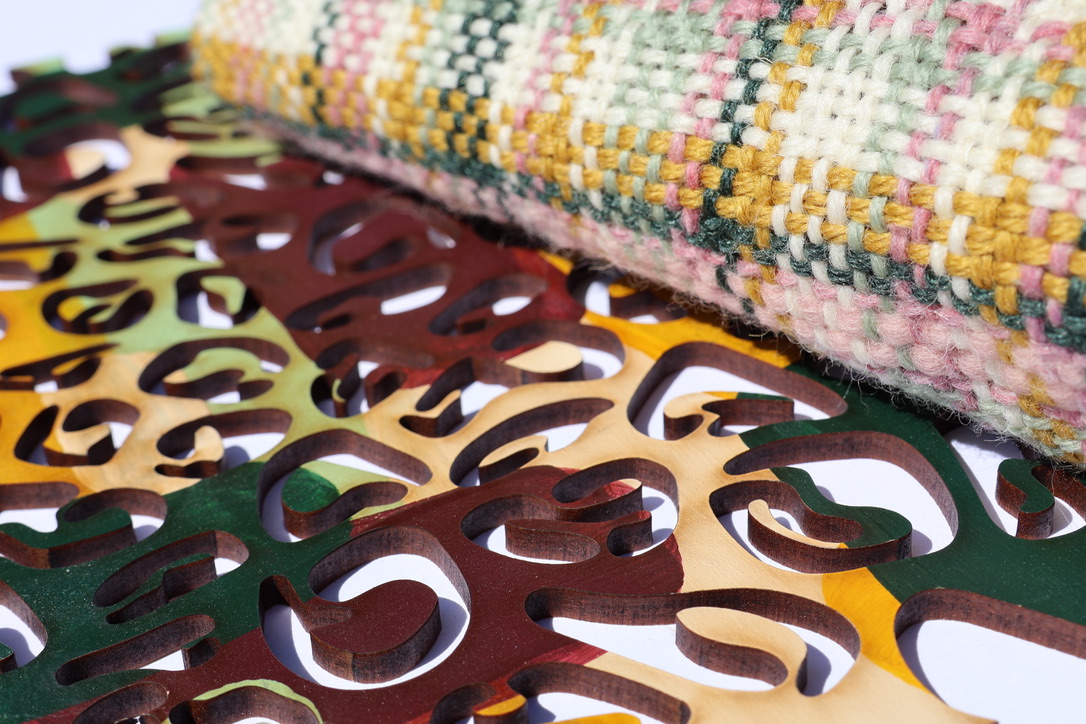 A textured woven fabric in muted colours lies on top of an intricately cut decorative overlay featuring various shapes in earthy tones. The background is plain, enhancing the contrast between the two layers.
