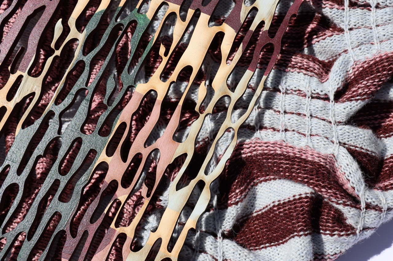 An abstract composition featuring several layered metal patterns in various metallic hues, including copper and bronze, against a textured background of knitted fabric in maroon and cream stripes.
