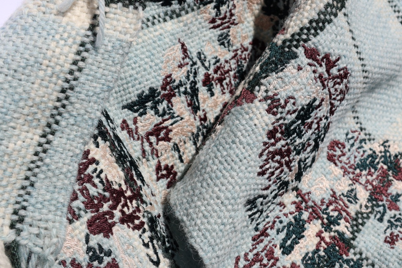 A close-up of a woven fabric featuring a floral design in shades of burgundy, blue, and cream. The intricate patterns blend together, showcasing the texture and craftsmanship of the textile.