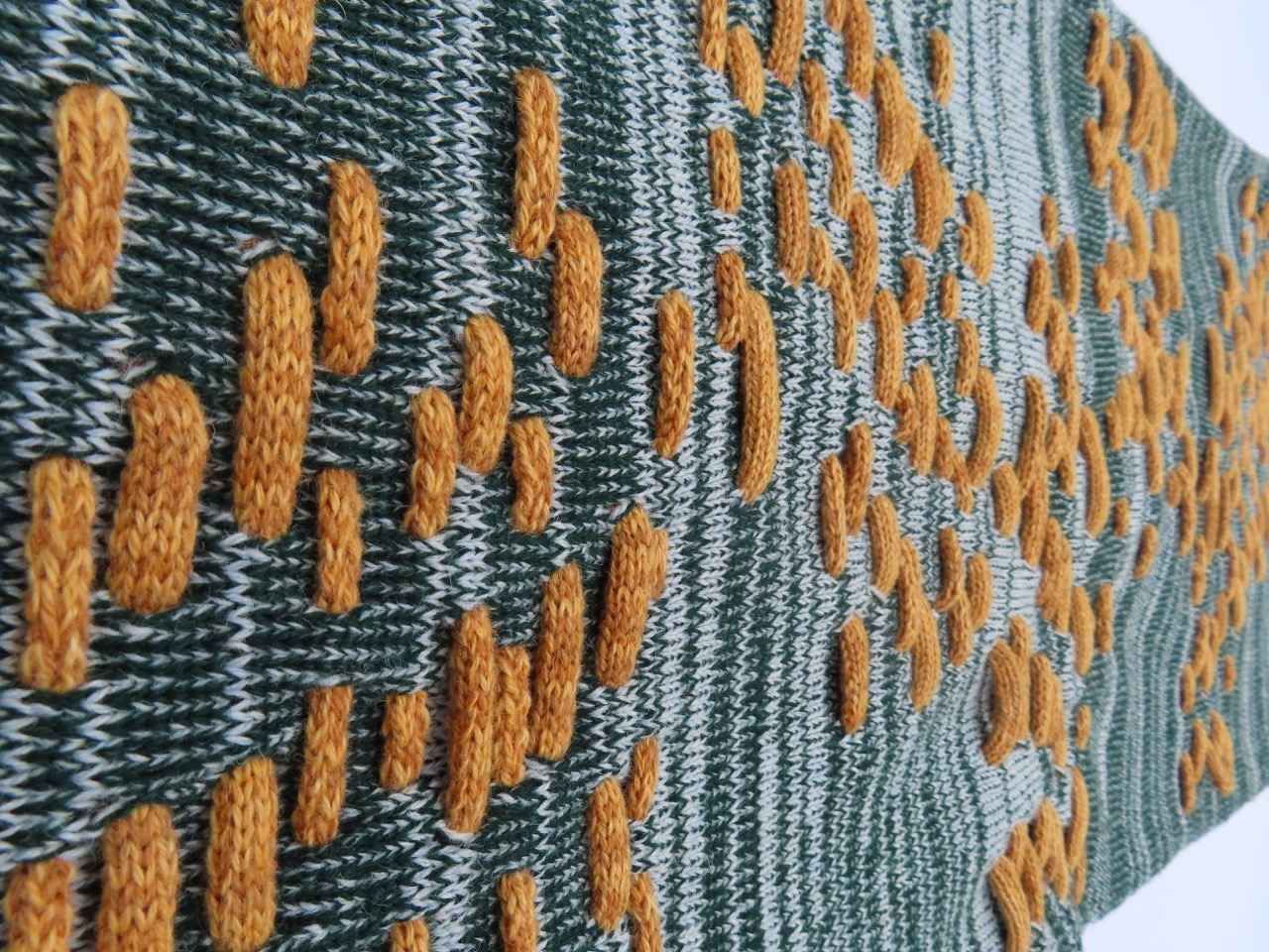 A knitted textile featuring a pattern of orange blobs on a green backdrop. The design shows varying sizes and orientations of the blobs, creating a textured appearance across the surface.