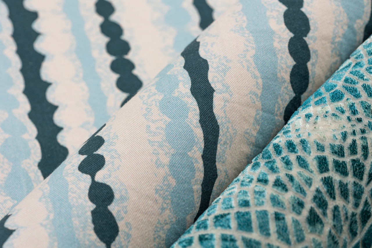 Close-up of three folded fabric swatches featuring patterns. The left swatch has a light blue and white wavy design, the middle shows a similar scheme with dark blue accents, and the right swatch displays a textured teal print with a grid-like pattern.