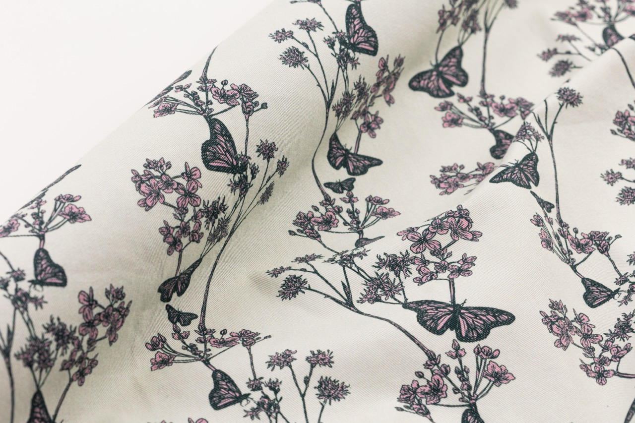 A close-up of patterned fabric featuring illustrations of butterflies and flowers. The design includes pink flowers and dark green outlines against a light background.