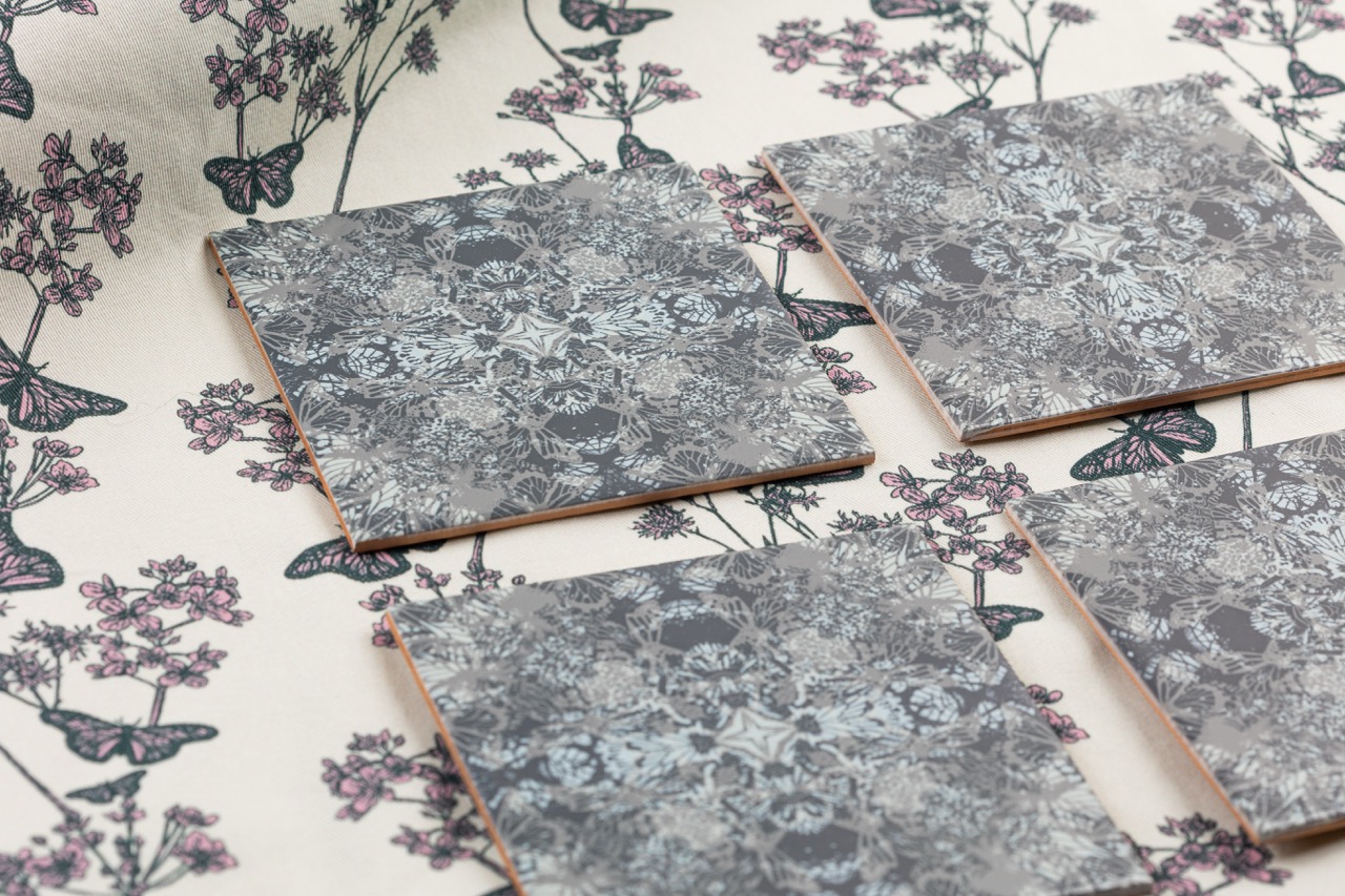 Four square ceramic tiles with a floral grey pattern are laid on a fabric surface featuring a similar floral design in muted colours. The background includes butterflies and delicate flowers, creating a cohesive design.