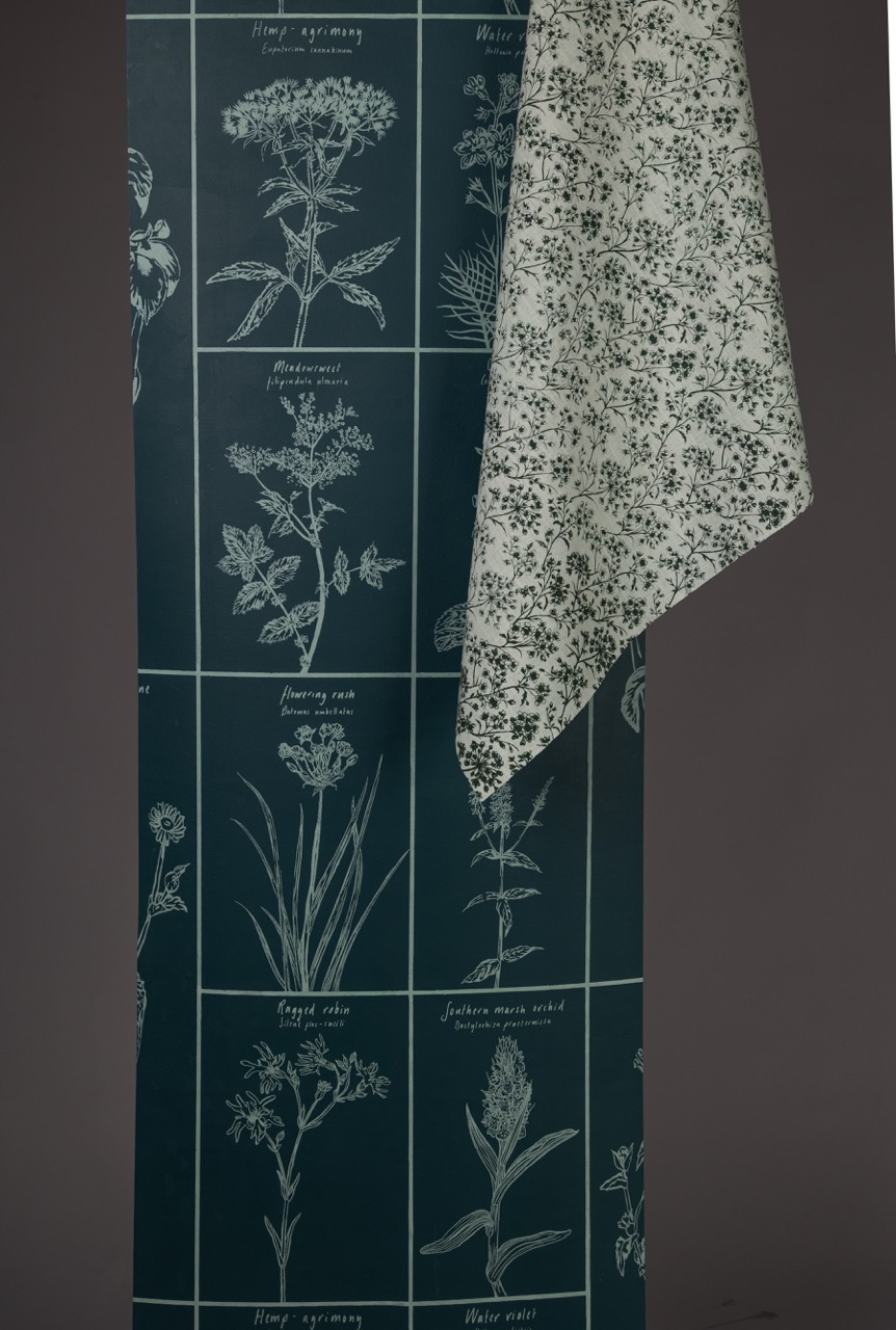 A dark green wallpaper featuring botanical illustrations of various plants, accompanied by a light floral-patterned fabric draped over one edge. The wallpaper has a grid layout with detailed sketches of herbs and flowers.