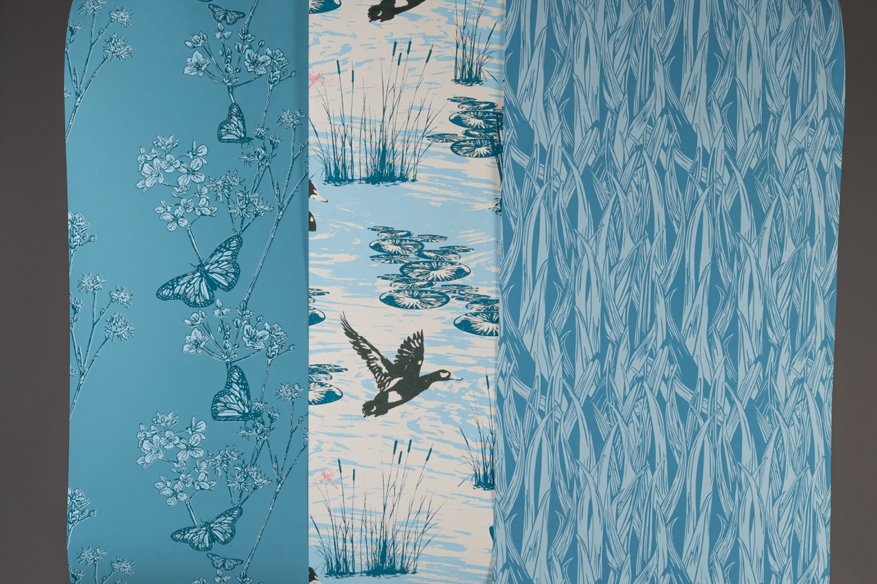 Three pieces of fabric displayed side by side. The left fabric is light blue with a floral and butterfly pattern, the middle features a water lily scene with ducks, and the right displays a stripe design resembling tall grasses in varying shades of blue.