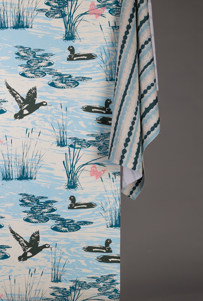A patterned fabric featuring water lilies, blue water, and ducks, along with green reeds and flowers. The design is complemented by an adjacent piece of striped fabric in shades of blue and white, partially visible.