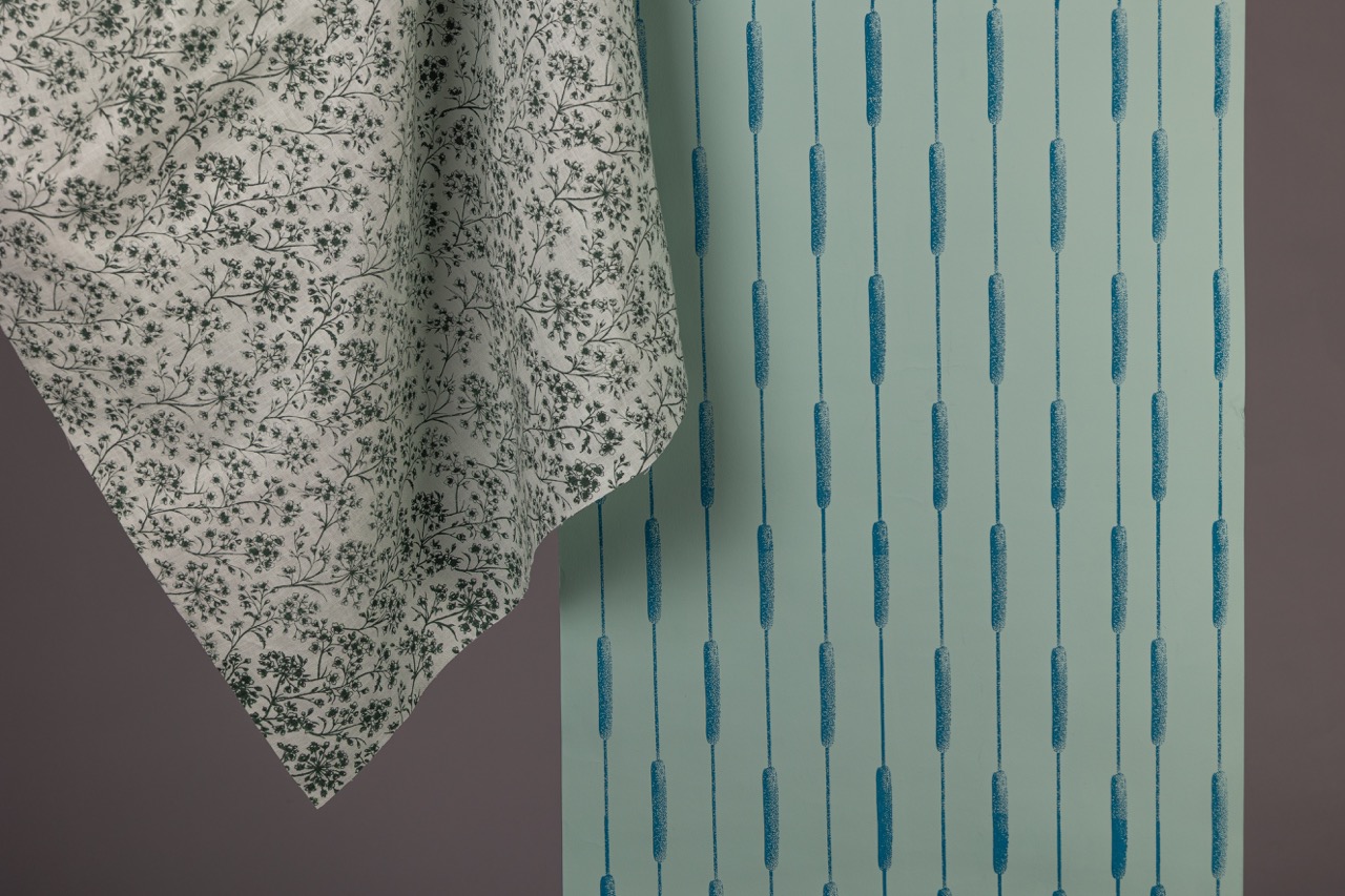 A white tablecloth with a green floral pattern drapes over the edge of a surface. Beside it, a blue wallpaper features vertical lines with a repeated design of stylised grass or plant forms, creating a contrasting visual texture.