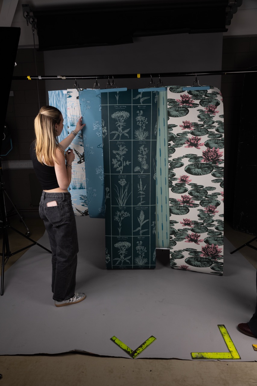 A woman with long blonde hair, wearing a black tank top and dark trousers, examines fabric samples hanging from a rack in a studio. The samples feature botanical designs on a light blue background and large floral patterns in greens and pinks.