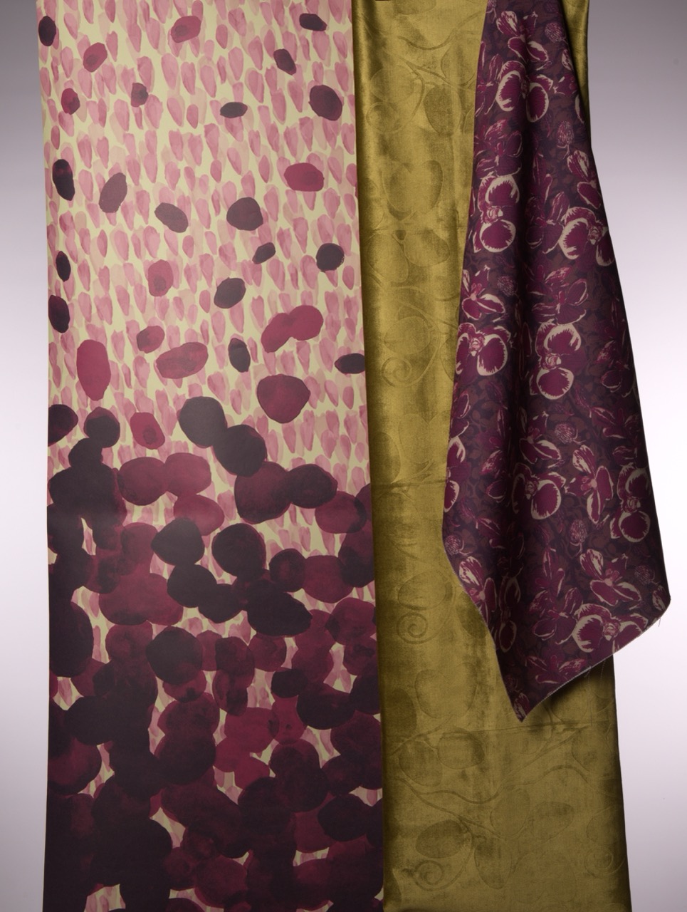 Three pieces of fabric are displayed. The left fabric features a floral pattern in shades of burgundy and cream, the centre is a solid olive green, and the right is a patterned deep purple fabric with swirling designs. The background is softly lit.