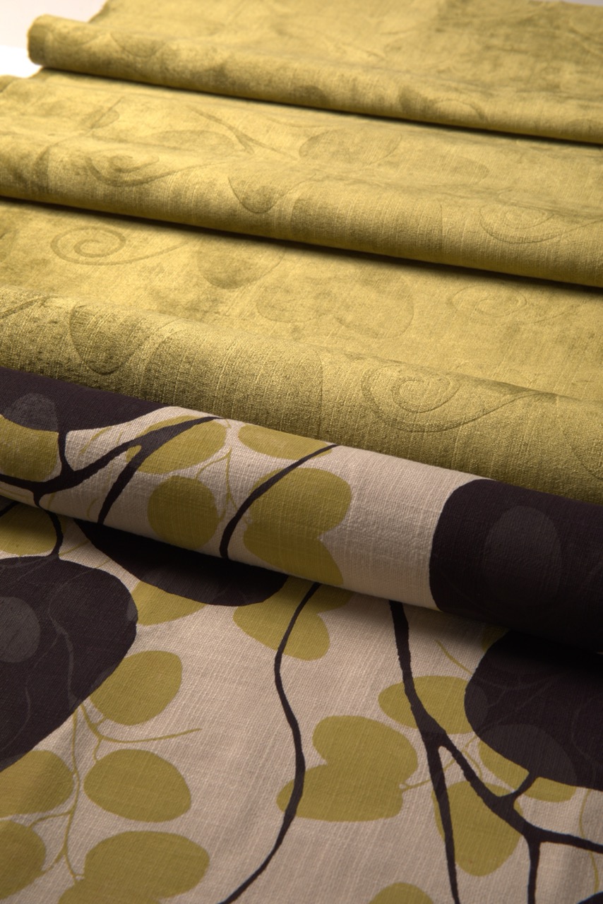 Folded fabric sheets in varying shades of green and beige, showcasing a floral pattern in dark grey and light green. The texture of the fabric ranges from smooth to slightly textured, arranged neatly in a stacked manner.