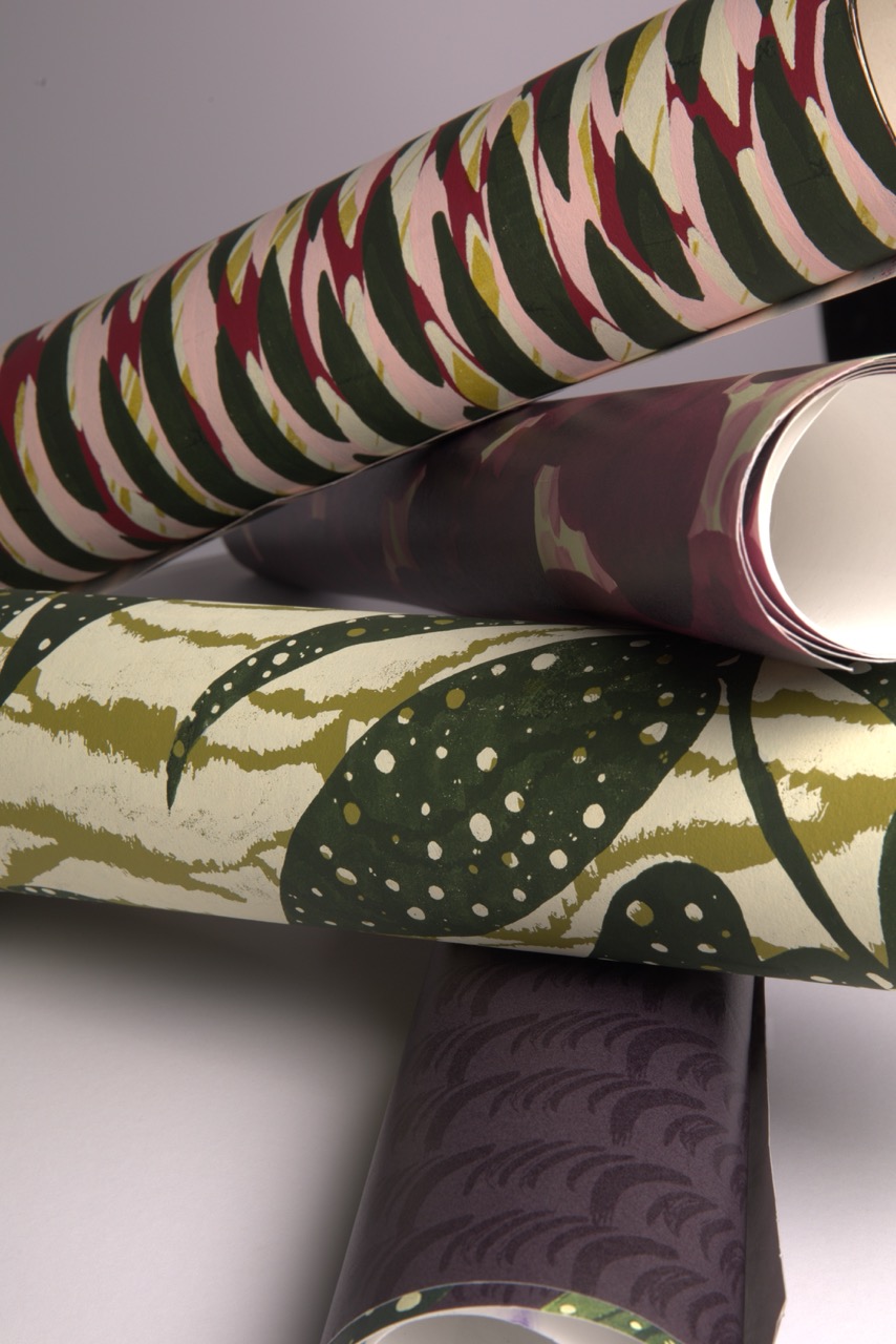 Three rolls of patterned wallpaper arranged in a stack. The top roll has a pink and green zigzag design, the middle roll features a dark maroon colour, and the bottom roll displays a green leaf pattern on a light background.