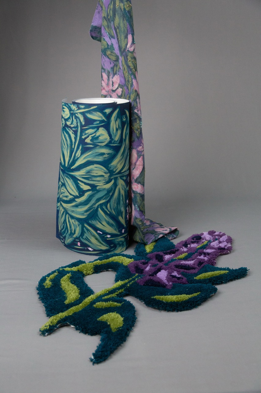 A cylindrical lamp with a blue-green leafy design stands beside a vibrant floral-patterned scarf, both placed on a grey surface. In front, a shaggy rug features a purple flower with green leaves, creating a cohesive botanical theme.