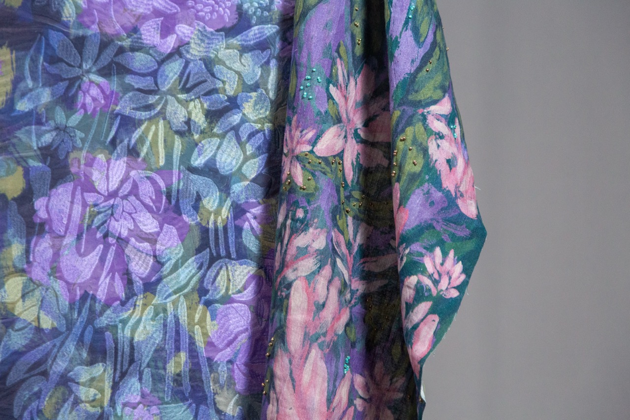 A close-up view of a colourful scarf featuring floral patterns in shades of purple, pink, and green. The fabric has a soft texture, with intricate designs blending seamlessly, creating a vibrant visual effect against a grey background.